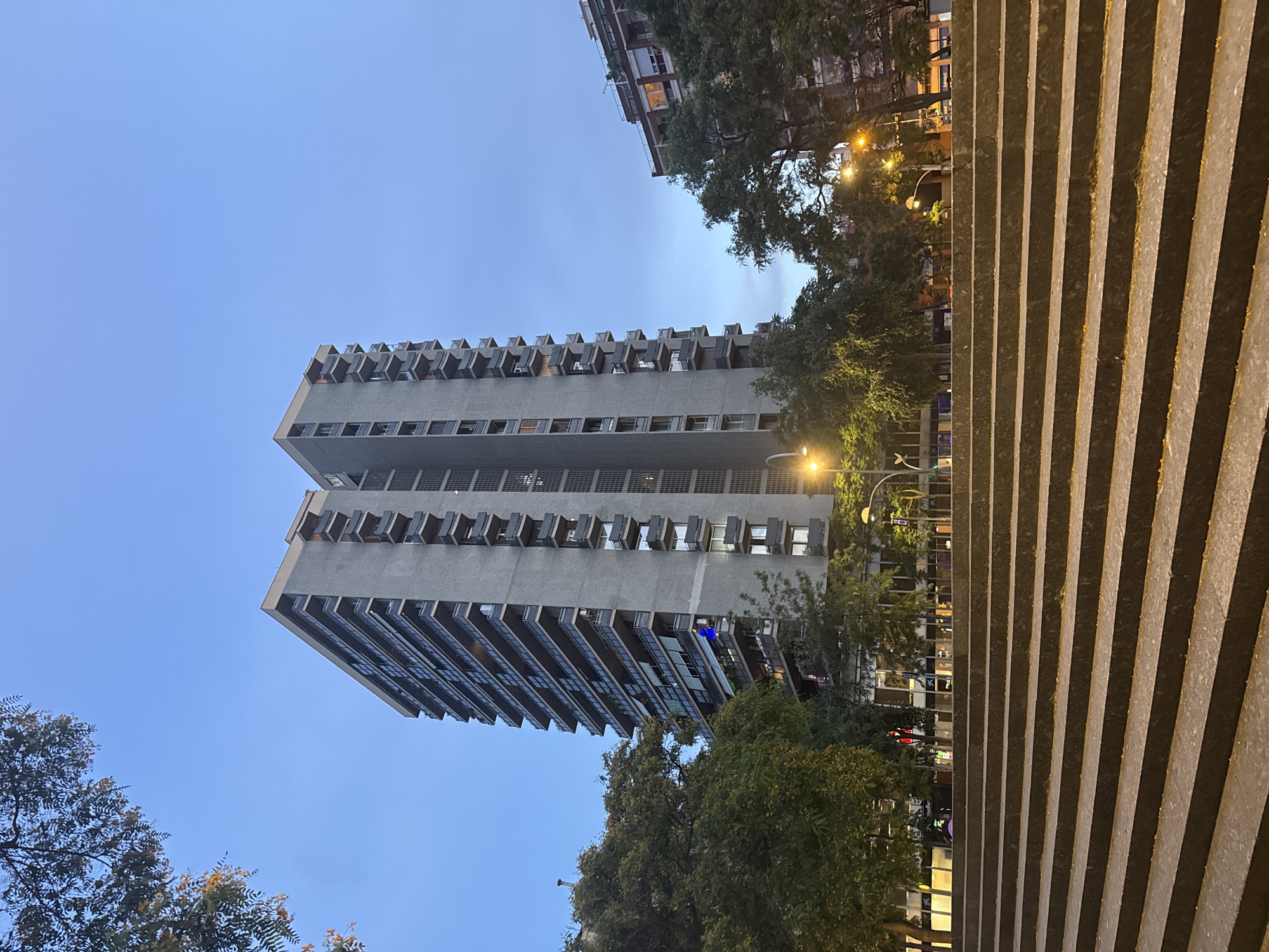 The image shows a tall, grey apartment building in Barcelona, Spain, at dusk. The building has many balconies with dark railings and glass fronts. Green trees with some yellowing leaves surround the base of the building. In the foreground, there is a wide set of stone stairs leading up to a paved area. Streetlights and lights from other buildings are visible, casting a warm glow on the scene. The sky is a deep blue, transitioning to a lighter shade at the horizon. There are no people visible in the image. The architecture suggests a modern urban environment.