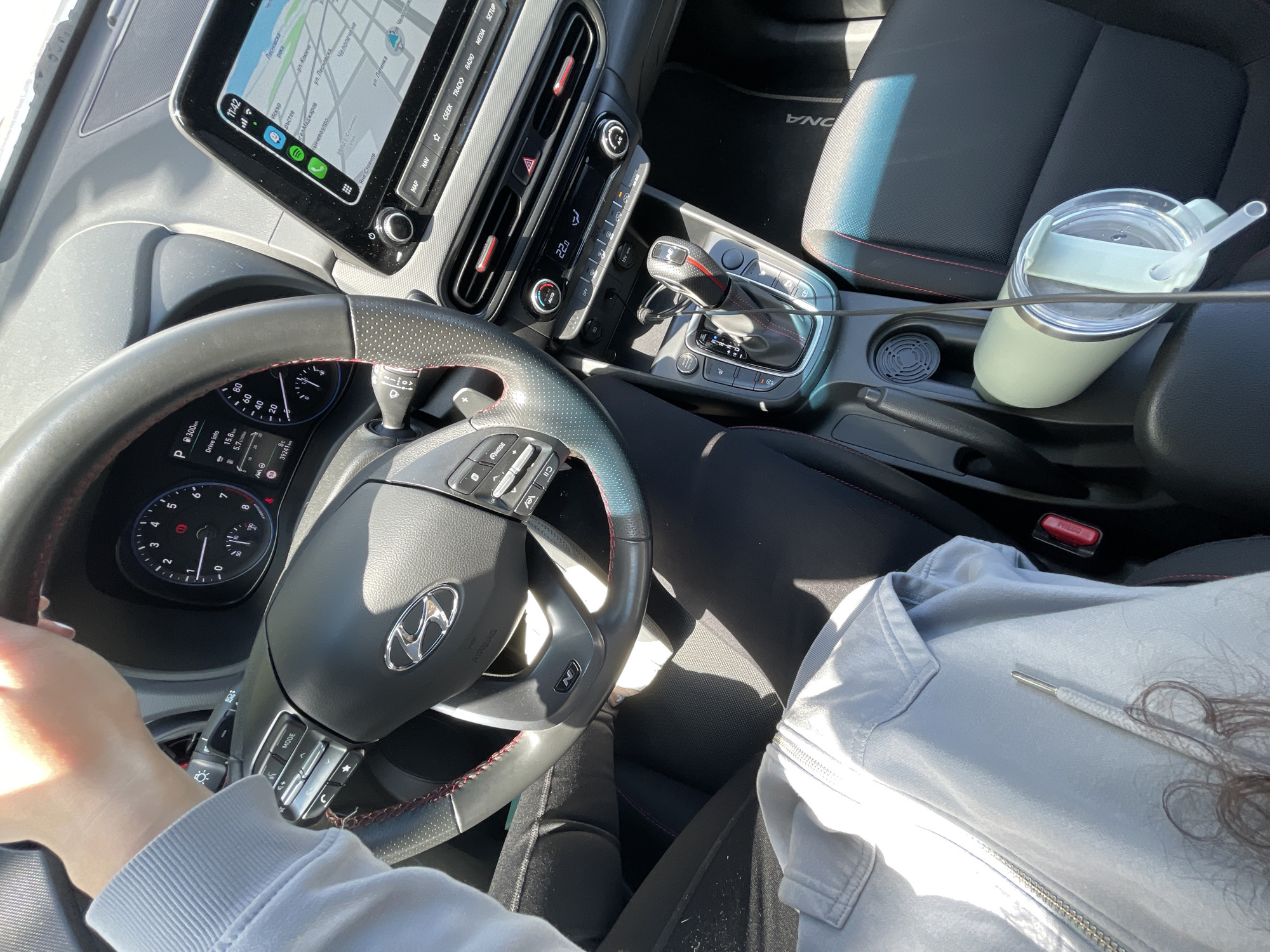 The image shows the interior of a car, specifically the driver's seat and dashboard area. A person's hands are visible on the steering wheel, and they are wearing a light grey jacket. The car's dashboard displays a navigation system showing a map of Sofia, Bulgaria. Place names visible on the screen are: Искър (Iskar), Кръстев (Krastev), Ангел Маджаров (Angel Madzharov), Слънчево утро (Slanchevo utro), Лесновска река (Lesnovska reka), Челопечене (Chelopechene). The car has a cup holder with a mint-colored tumbler with a straw. The gear shift is in the center console. The seat is black with red stitching.