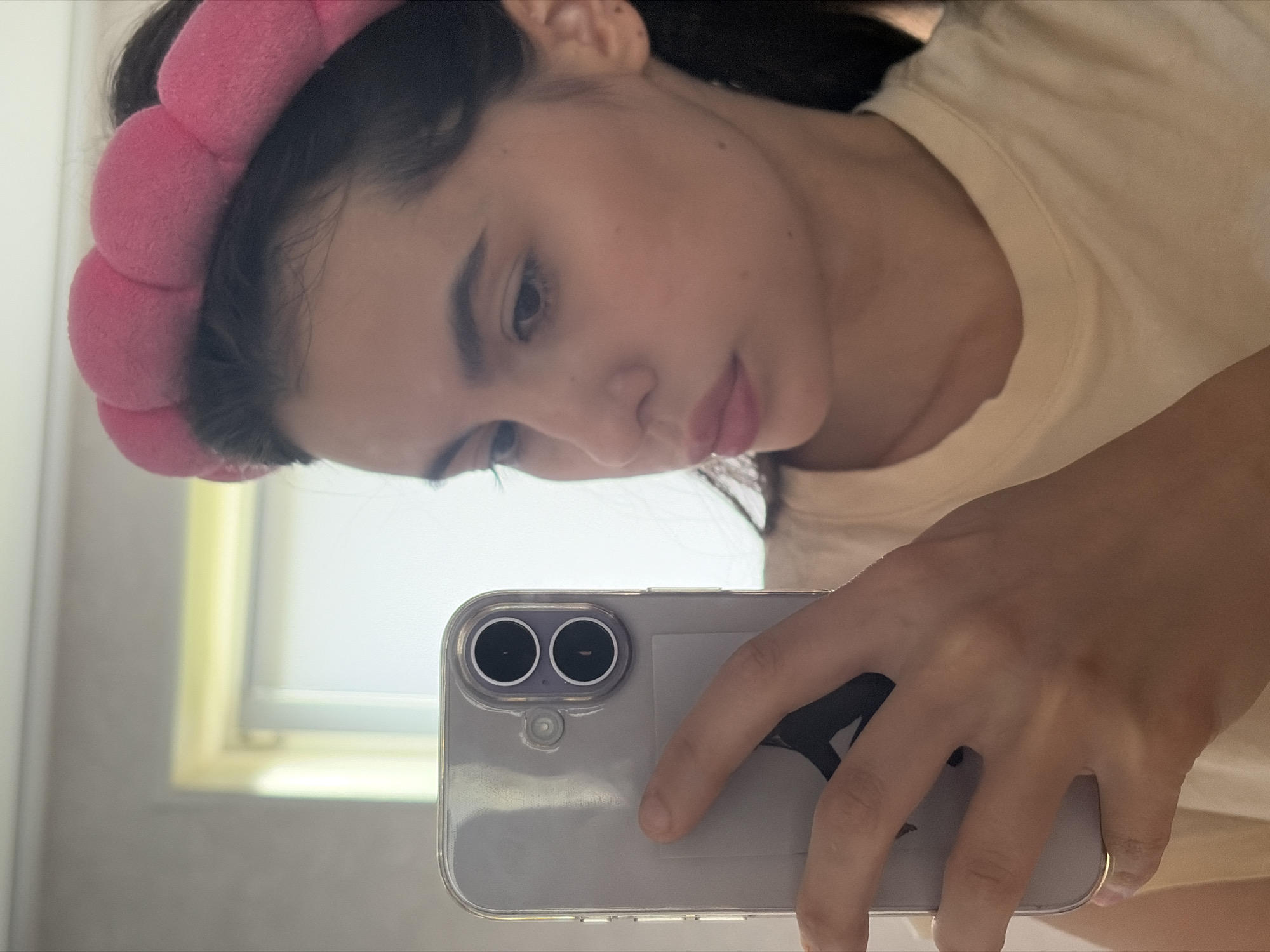 A young woman, likely in her late teens or early twenties, is taking a selfie in a bathroom. She is wearing a fluffy pink headband and a light-colored t-shirt. Her hair is dark and pulled back. She is holding a clear phone case with a visible camera module and flash. The background shows a lightly textured wall and a window with natural light. There is no indication of this scene being in Chaikovsky, Russia.