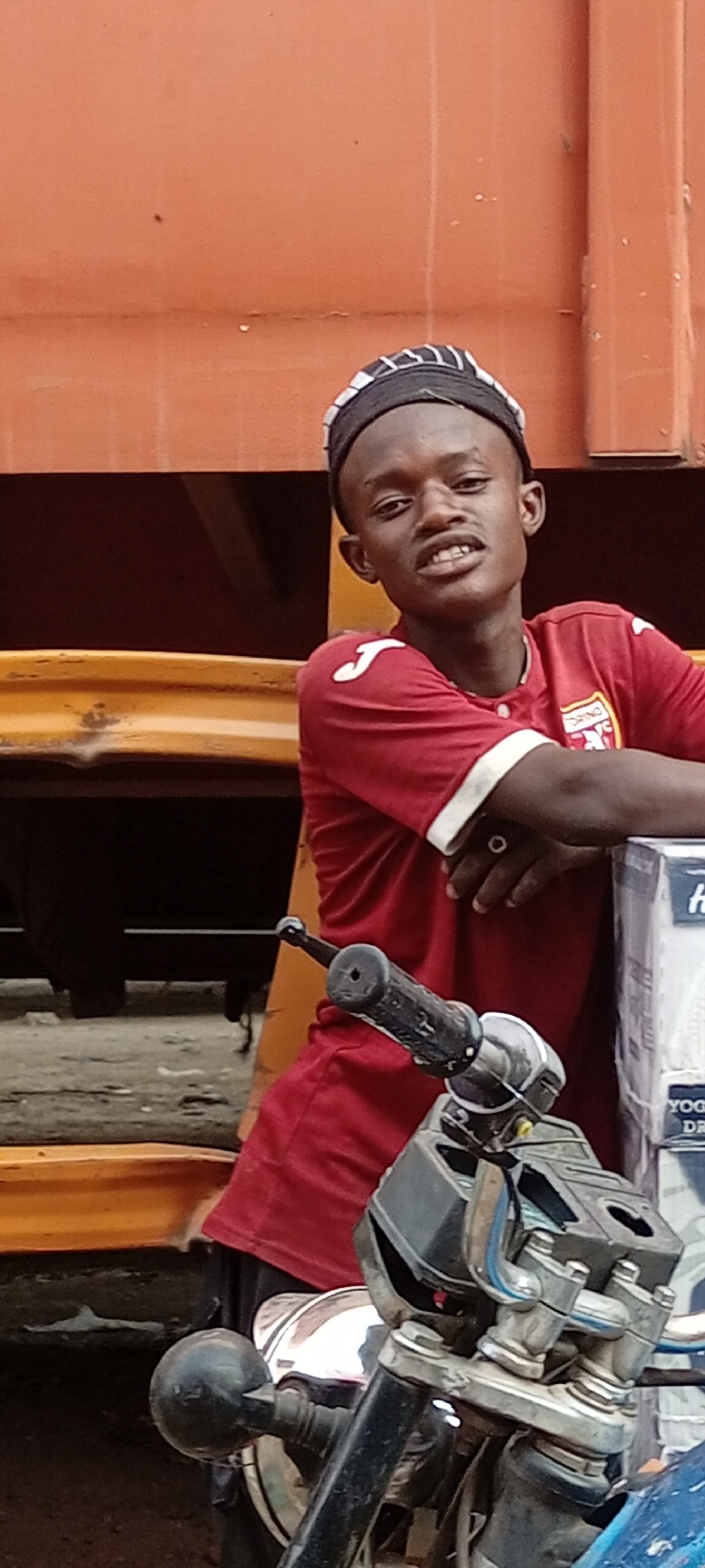 A young man wearing a burgundy Torino FC soccer jersey, a black and white striped hat, and a ring on his left hand stands in front of a yellow truck. He is leaning on a box that appears to be on a motorcycle. The location is Fagge A, Nigeria, evidenced by visual markers and common features of Nigerian urban scenes.