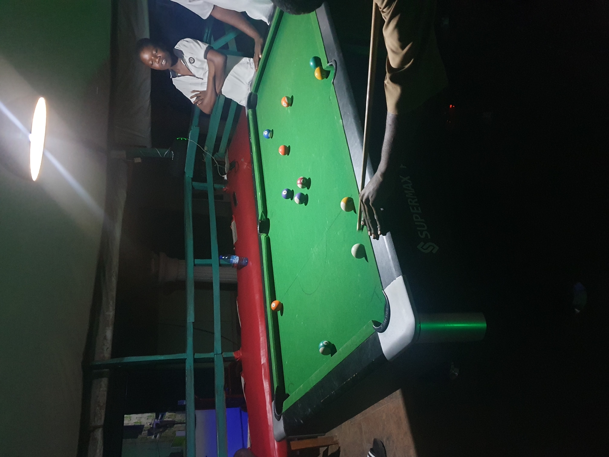 The image shows an indoor pool hall at night, likely in Lokoja, Nigeria, based on the context. A green pool table with several balls is central to the scene. Two young men are visible, one standing with arms crossed, wearing a white polo shirt, and another partially visible, holding a pool cue and appearing to play. The environment is dimly lit, with a bright overhead light illuminating the pool table and a portion of the ceiling. The background suggests an outdoor setting with some roofing and railings, possibly a bar or recreational area.