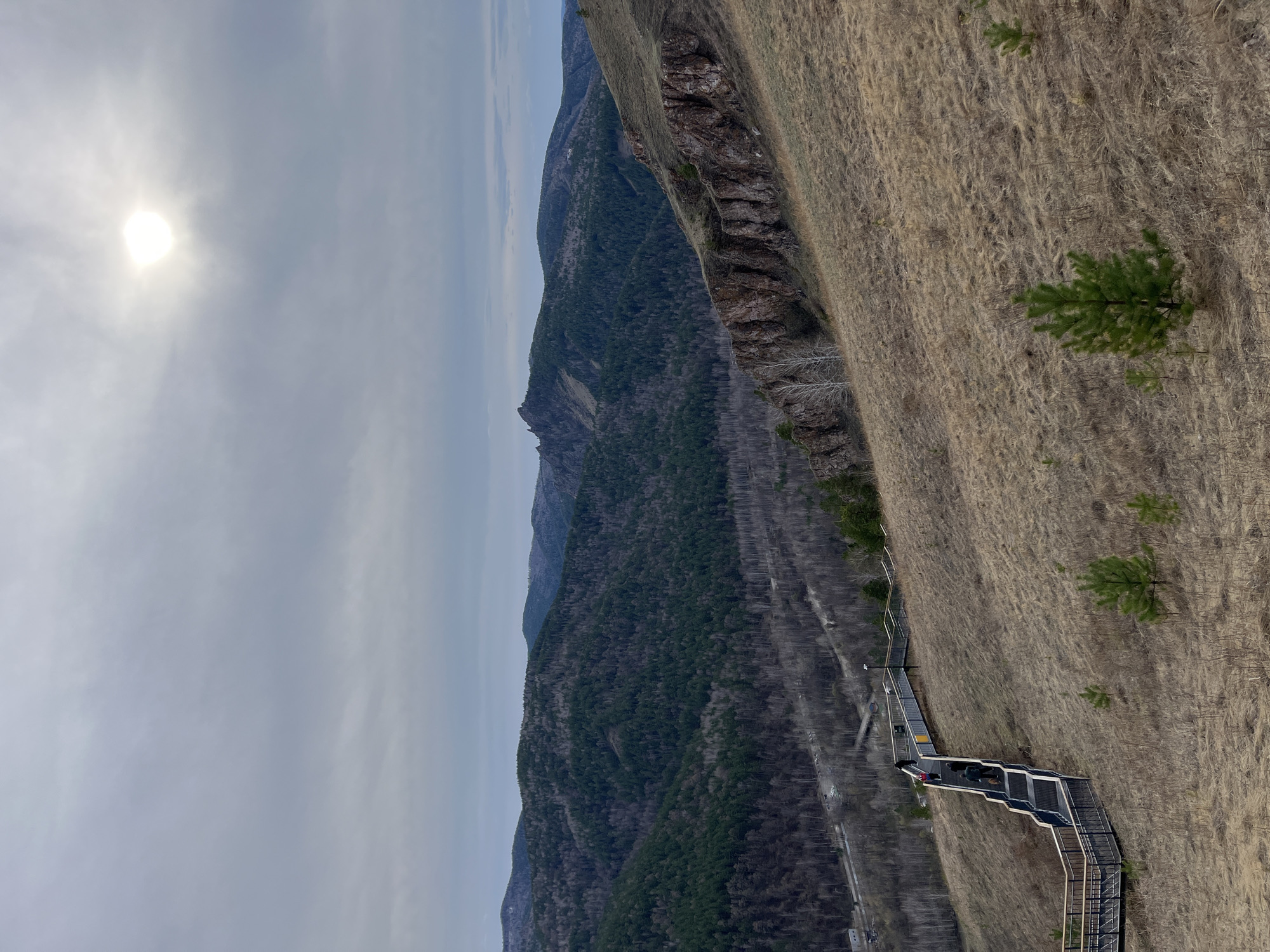 The image captures a view of a mountain landscape in Krasnoyarsk, Russia. The sun shines faintly through a cloudy sky. A mountain range is visible in the distance, covered in dense forest. In the foreground, a steep slope of dry, grassy terrain descends to a walking path, with a metal stairway descending down the hill, with figures visible walking on the stairs. The landscape appears rugged and natural.