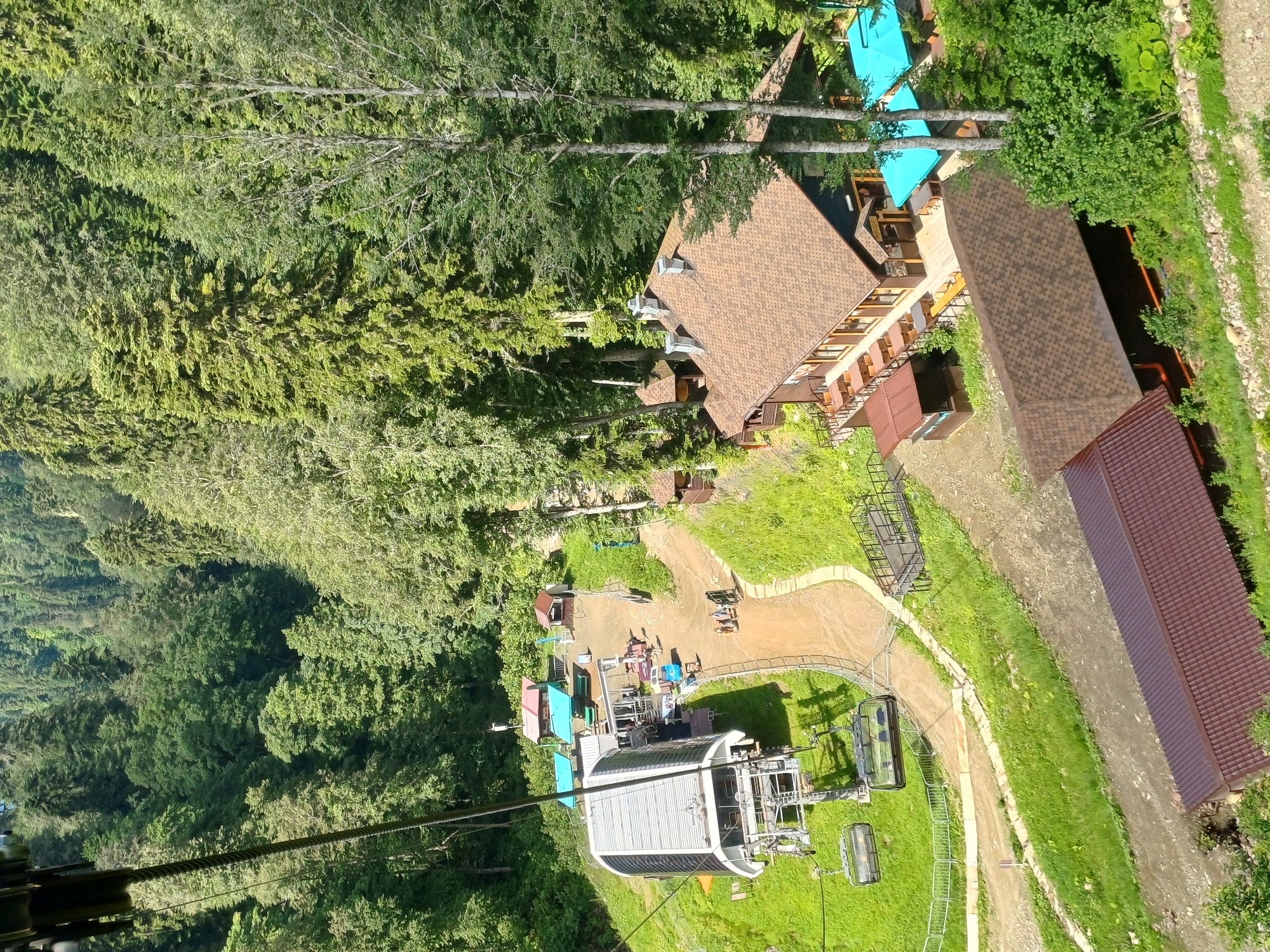 This image depicts a view from a ski lift in Estosadok, Russia, overlooking a mountain resort area. In the foreground, two ski lift chairs are suspended from a cable, implying the viewpoint is from a moving lift. Below, a modern ski lift station with a large, silver roofed building is visible, with several ski lift chairs docked. A dirt path winds around the station, leading to a cluster of buildings. One building is a two-story restaurant with a brown shingled roof and outdoor seating areas with blue umbrellas. Adjacent to the restaurant is a smaller building with a similar roof. Further down the path, there are picnic tables with colorful awnings and a small kiosk. Several people are visible near the ski lift station and on the path, appearing to be tourists or resort staff. The surrounding environment is a dense, green forest with tall evergreen trees, typical of a mountain region. The overall scene conveys a sense of outdoor recreation and a functional resort setting within a natural landscape.