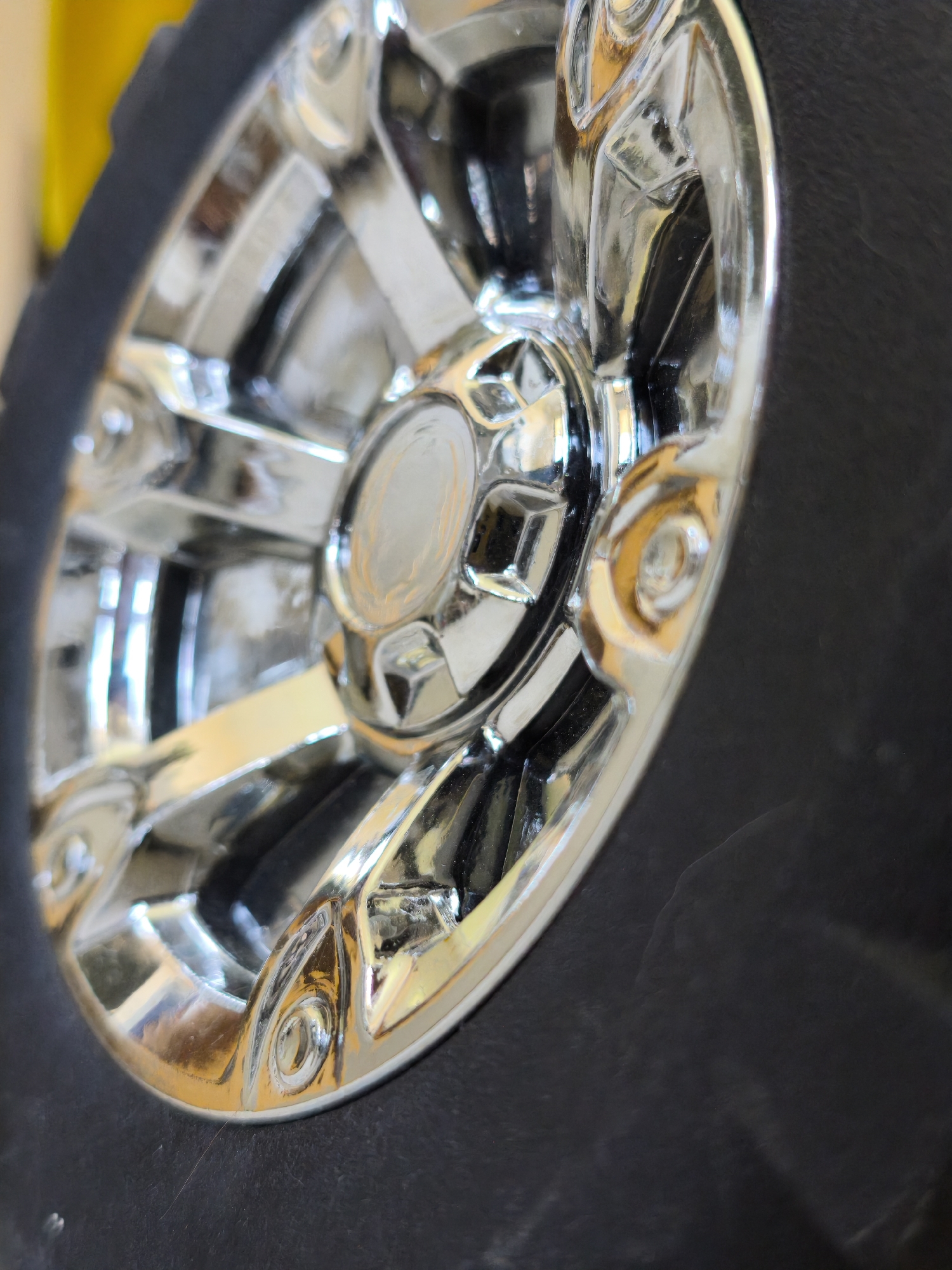 A close-up shot of a tire with a chrome-finished wheel. The tire is black, and the wheel has a multi-spoke design with a circular hub. The chrome surface reflects light, creating highlights and shadows. Part of a yellow object appears in the background. There is no evidence this image was taken in Plovdiv, Bulgaria.
