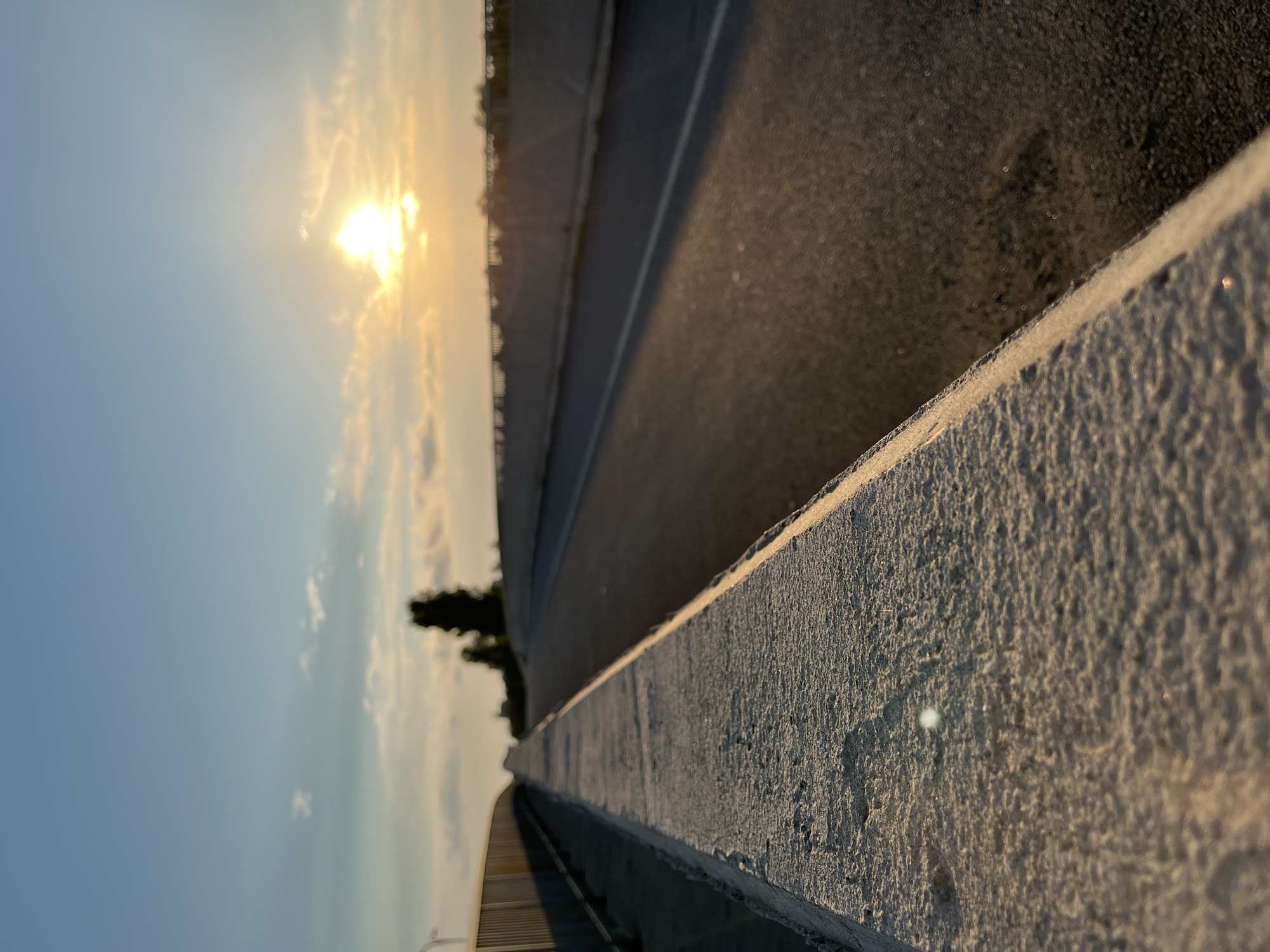 The image depicts a perspective view of a road on a bridge at sunset. The foreground features a rough-textured concrete barrier on the left, with the road extending into the background on the right. The road is asphalt with a white line marking its edge. The setting sun is visible in the upper right quadrant of the image, casting a warm glow and creating lens flare. Two silhouetted trees stand in the distance, behind a concrete barrier on the right side of the road. The sky is a gradient of blue and orange, with scattered clouds. The overall scene is serene, capturing the atmosphere of a bridge overlooking a landscape at dusk. The location is specified as the city of Ureki, Georgia.
