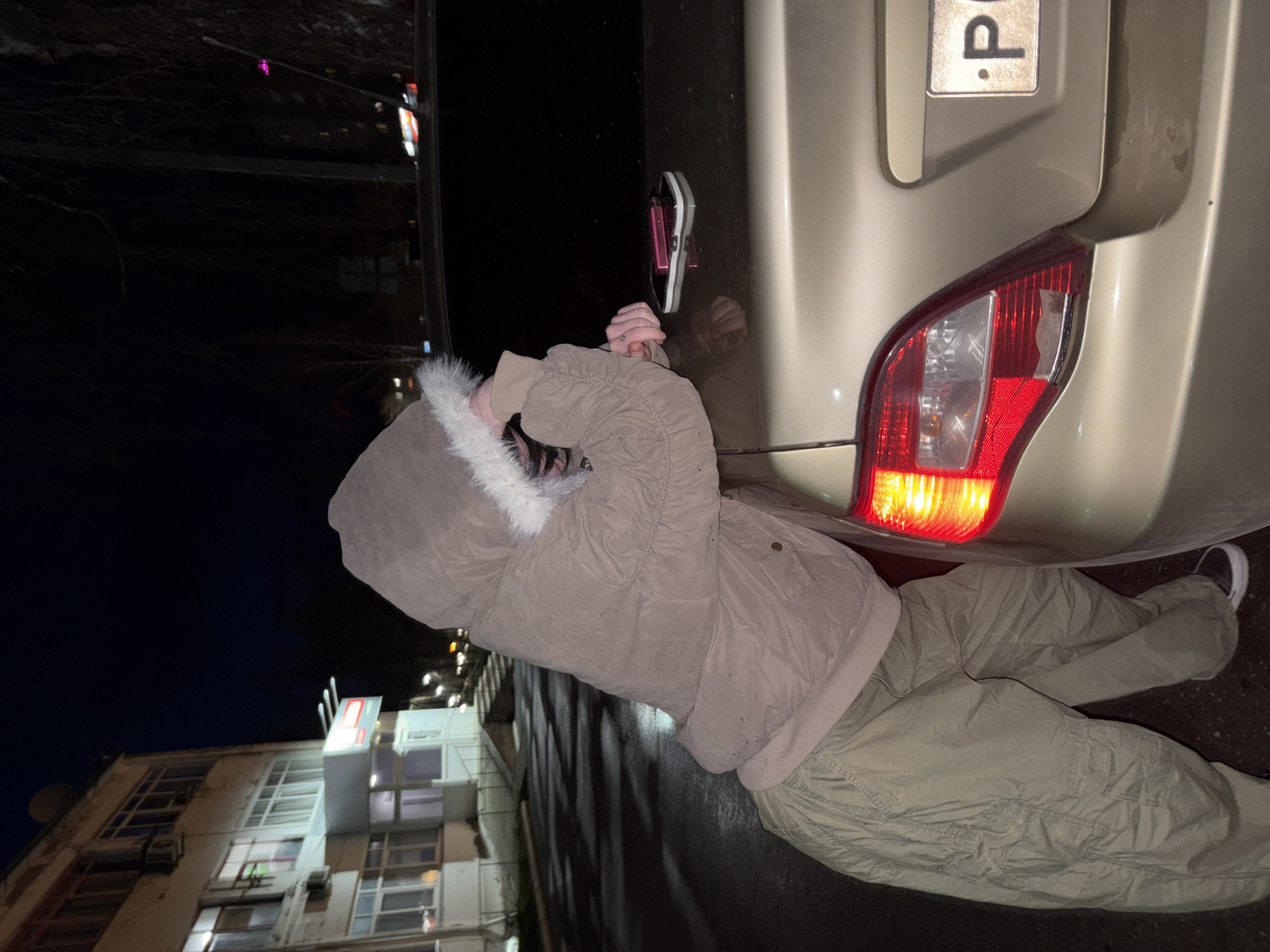 An outdoor, eye-level shot at night in Chaikovsky, Russia, shows a person inspecting the rear of a light-colored (beige/gold) car. The person, viewed from behind, wears a beige/tan hooded winter jacket with white fur trim and matching cargo pants. They are bent over, with their hands near the car's trunk area.
A notable detail is a pink and white shoe or sandal, with a black sole, seemingly attached to the car's rear quarter panel/door, near the person's hands. The car's right taillight is illuminated, suggesting it's on, and appears to have a broken or damaged cover. Above the taillight, a partial Russian license plate is visible, showing "P.".
The setting is dark, illuminated by artificial light sources. In the background, a multi-story building with numerous windows and what appear to be air conditioning units is visible. Bare tree branches are silhouetted against the dark sky. The ground beneath the car is dark and appears to be pavement or asphalt. The person's winter attire suggests cold weather. The image captures a moment of focused attention on the car's rear, particularly the attached shoe and the damaged taillight.