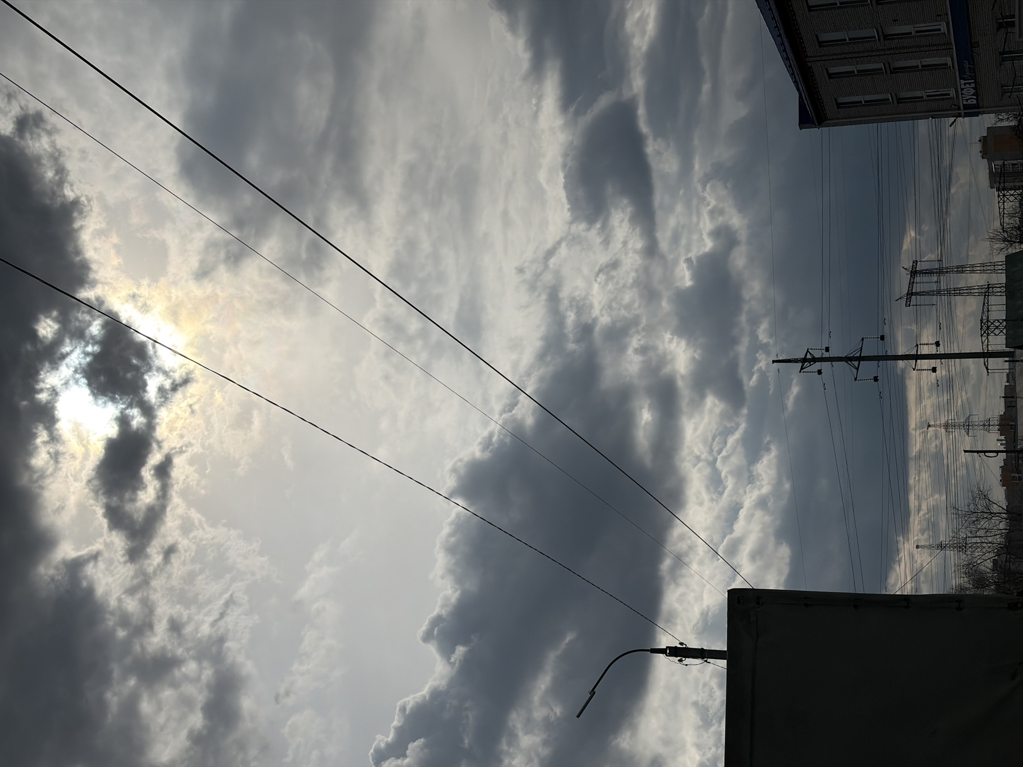The image depicts an urban or suburban landscape dominated by a dramatic, overcast sky. Dark grey and lighter white clouds fill the frame, with the sun partially breaking through in the upper left quadrant, creating a bright, glowing patch of sky. Several dark power lines stretch diagonally across the sky.
On the right side of the image, the top section of a multi-story brick building is visible, featuring multiple windows and a dark blue band along its roofline. On this blue band, the Russian word "БУФЕТ" (Bufet, meaning 'buffet' or 'canteen') is clearly visible in white capital letters. Below and to the right of this building, a series of tall utility poles and electricity transmission towers are silhouetted against the lighter parts of the sky, with numerous wires running between them. Bare tree branches are discernible near the base of these structures.
In the lower left foreground, a street light fixture with a curved arm is seen, and below it, a large, dark, rectangular object, possibly a utility box or a large sign, occupies the bottom corner. The scene captures a daytime moment under a heavily clouded sky, likely in an urban area of Chaikovsky, Russia, as indicated by the specific infrastructure and visible text.
