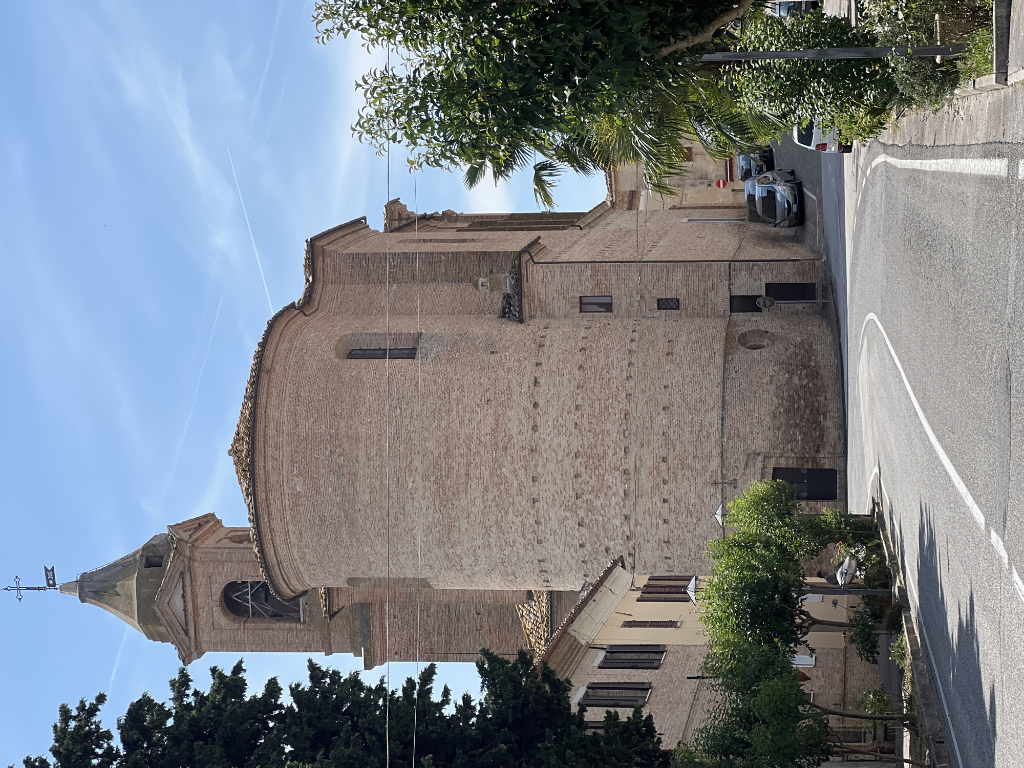 In the city of Barbara, Italy, on a sunny day with a clear blue sky, a brick church with a bell tower dominates the scene. The church's rounded apse faces a curving asphalt road with white lane markings. On the left, a tree casts a shadow on the road, partially obscuring a building with closed shutters. A few parked cars are visible further down the road, and on the right, lush green trees and bushes line the sidewalk. The scene captures a quiet moment in the Italian town, with the historic architecture and natural elements creating a picturesque atmosphere.