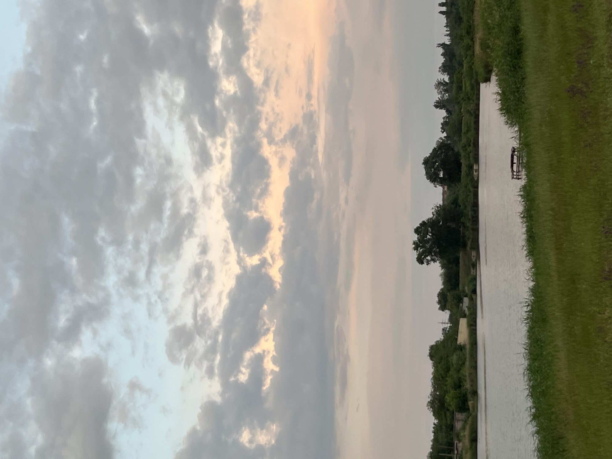 This is a photograph taken in Kryvyi Rih, Ukraine, depicting a scenic view of a body of water, likely a river or lake, with a lush green embankment in the foreground. The water reflects the muted colors of the sky, which is predominantly filled with soft, grey clouds tinged with hints of pale blue and a subtle peachy hue, suggesting it might be late afternoon or early evening.
The embankment is covered in dense, tall green grass, with some patches of purple wildflowers visible. Beyond the grassy area, the water stretches out, appearing calm and still. On the far side of the water, a line of trees and bushes forms a verdant backdrop. Amongst the foliage, there are some structures visible, including what appears to be a light-colored building and possibly some industrial elements in the distance, hinting at the proximity of urban development within the otherwise natural landscape.
In the water, near the right side of the frame, a small wooden platform or pier is visible, extending a short distance from the shore, suggesting a place for recreation or access to the water. The overall atmosphere of the image is tranquil and serene, with the natural beauty of the environment taking center stage under a dramatic sky. There are no people or animals visible in the photograph.