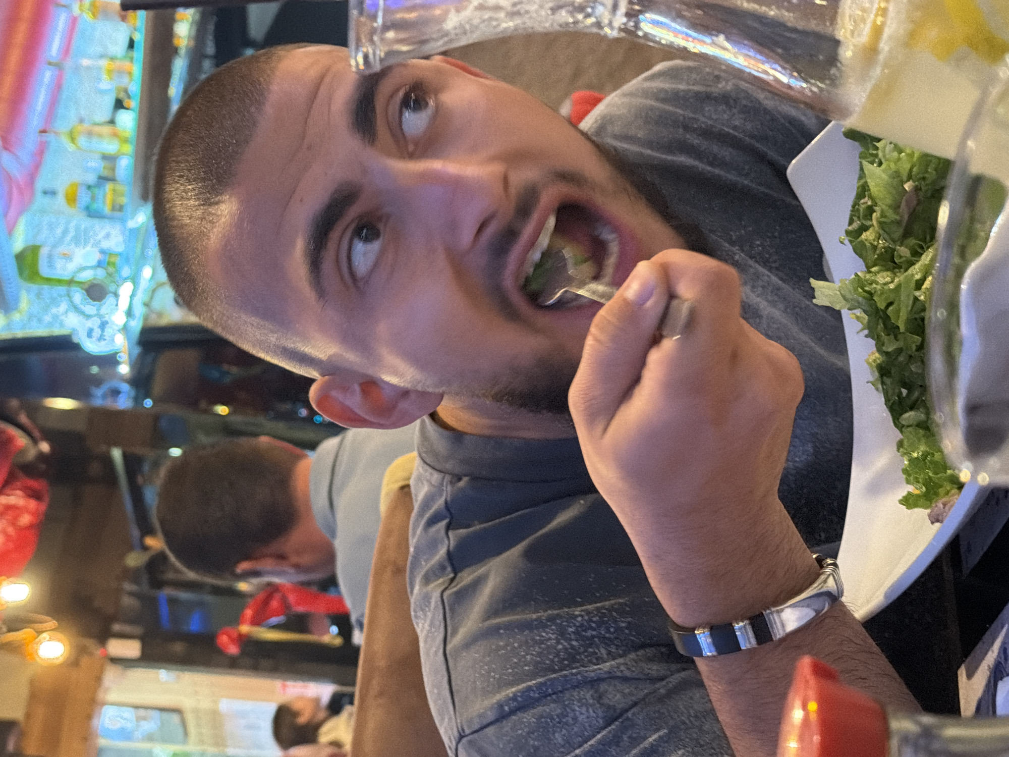 An image, rotated on its side, captures a man in the foreground actively eating a salad in an indoor setting, likely a restaurant or bar. The man has short, dark hair and stubble, wearing a dark grey t-shirt and a silver and black bracelet on his right wrist. His mouth is wide open as he brings a forkful of green salad to his mouth, and his eyes are looking upwards with a wide-eyed, expressive gaze, suggesting surprise or being caught mid-action.
To the right of the man is a white plate holding a substantial serving of green leaf lettuce. A clear glass or bottle is partially visible in the upper right, and another reflective object, possibly part of a drinkware, is seen above his head. The background is blurred but shows the distinct elements of a bar, including shelves filled with numerous glowing liquor bottles in various colors, creating a vibrant, illuminated effect. Another man with dark, short hair is partially visible further in the background, facing away from the camera and seemingly seated at a table. The overall lighting is dim, emphasizing the glowing bottles and the subjects in the foreground. No discernible text is visible on any objects or in the background. The time of day appears to be evening or night, given the artificial lighting and bar environment.