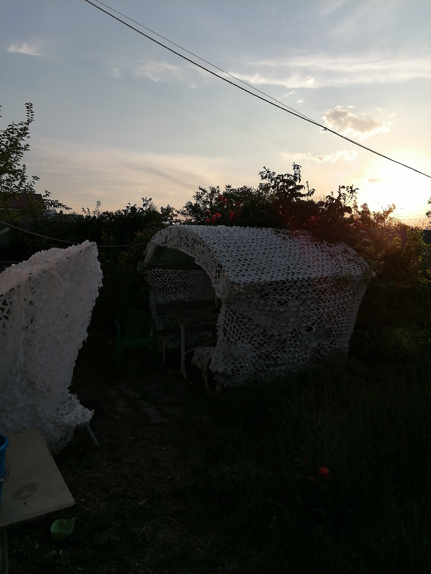 The image depicts a backyard scene at dusk, likely in Rostov-on-Don, Russia, given the context. The sky is illuminated by the setting sun, casting a warm glow through scattered clouds. In the foreground, to the left, is a white camouflage net draped over an unseen object, appearing somewhat like a partition. To the right, a larger, white camouflage net structure covers what appears to be an outdoor seating area, possibly a gazebo or a table with chairs. A green plastic chair is visible peeking out from under this structure. Behind the covered seating area, dense foliage and trees are silhouetted against the sky, with hints of red flowers amongst the greenery. The ground is covered in dark gravel or small stones. A power line stretches across the upper part of the frame. The overall impression is one of a quiet, domestic outdoor space transitioning into evening.