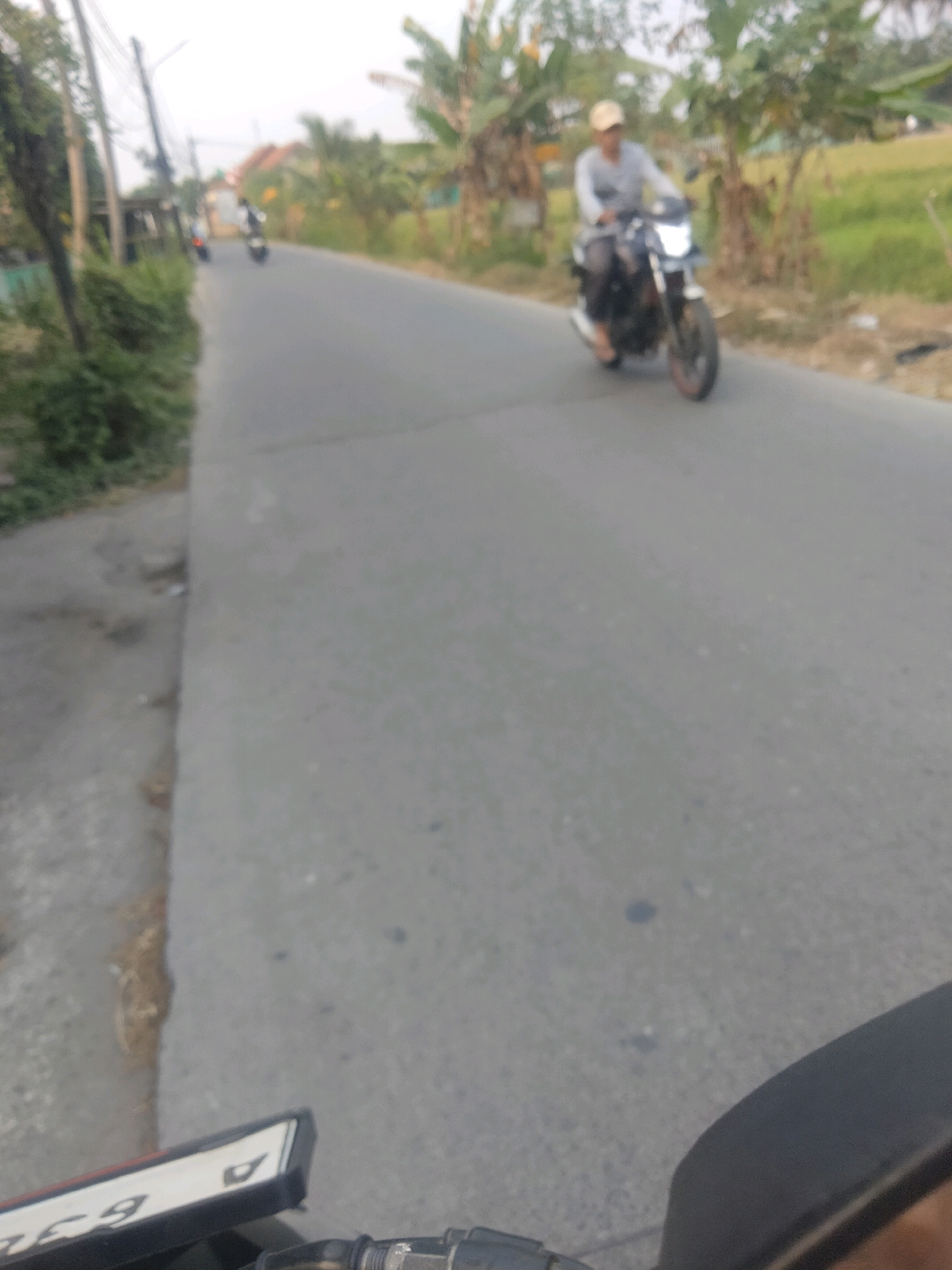 The image captures a street scene in Suka Sari, Indonesia, on a clear day. A person is riding a motorcycle down the asphalt road, which is flanked by vegetation and trees. The rider, wearing a light-colored shirt and a cap, is blurred due to motion. A second motorcycle is visible further down the road, also in motion. The foreground of the image shows a portion of another motorcycle's license plate, partially obscuring the view. The overall scene suggests a casual, everyday commute or travel within the village.