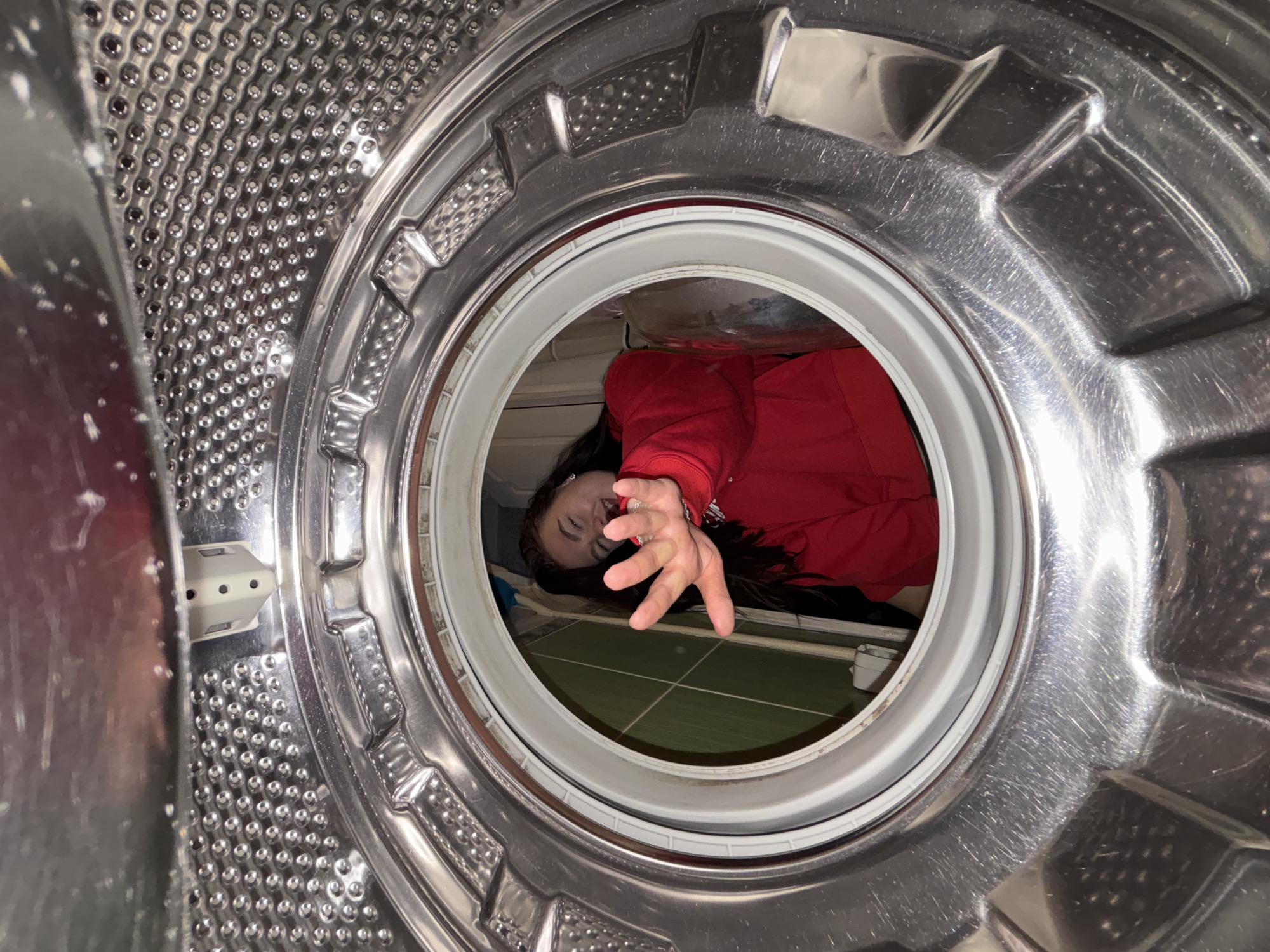 The image presents a unique, low-angle perspective from inside the metallic drum of a washing machine, looking outwards. The textured interior of the drum features a pattern of small, dimpled indentations and several larger, curved metal fins that form the inner frame. A clean white rubber seal encircles the circular opening of the washing machine.
Through this opening, a person is visible, positioned in front of the appliance. She appears to be a woman with long, dark hair, wearing a vibrant red long-sleeved top. Her head is tilted, and her right hand, adorned with a ring, is extended towards the washing machine's interior, as if reaching out to the camera or playfully interacting with the machine. Her expression seems to be a slight, engaged smile.
The background visible beyond the person reveals a room with a floor covered in square green and grey tiles, suggesting a laundry room or bathroom setting. Part of a light-colored object or piece of furniture is also visible behind her. The overall scene captures a candid, perhaps humorous, moment of a person engaging with a household appliance. The location context provided indicates this scene takes place in Perm, Russia. No discernible text is visible within the image.
