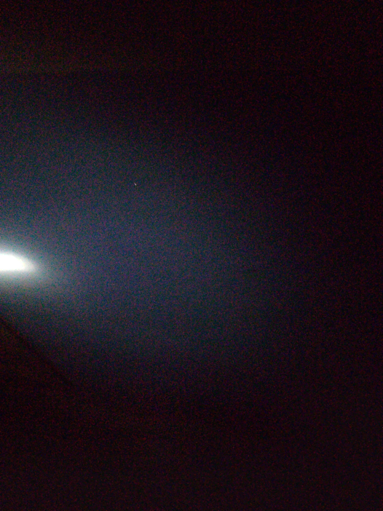 The image displays a dark, grainy scene with a bright light source on the left side. The light appears to be emanating from an artificial source and casts a faint glow across the left portion of the frame, with a subtle gradient of illumination. The rest of the image is predominantly dark, with subtle textures and noise visible throughout. There is a small, faint point of light in the upper right quadrant of the image, possibly a distant star or celestial body. No people, specific objects, or clear environmental features are discernible beyond the light source and the dark, textured background. The image's composition suggests a low-light or nighttime setting, possibly outdoors, but the lack of identifiable landmarks makes it impossible to determine a specific location within Jalingo, Nigeria, or any other city.