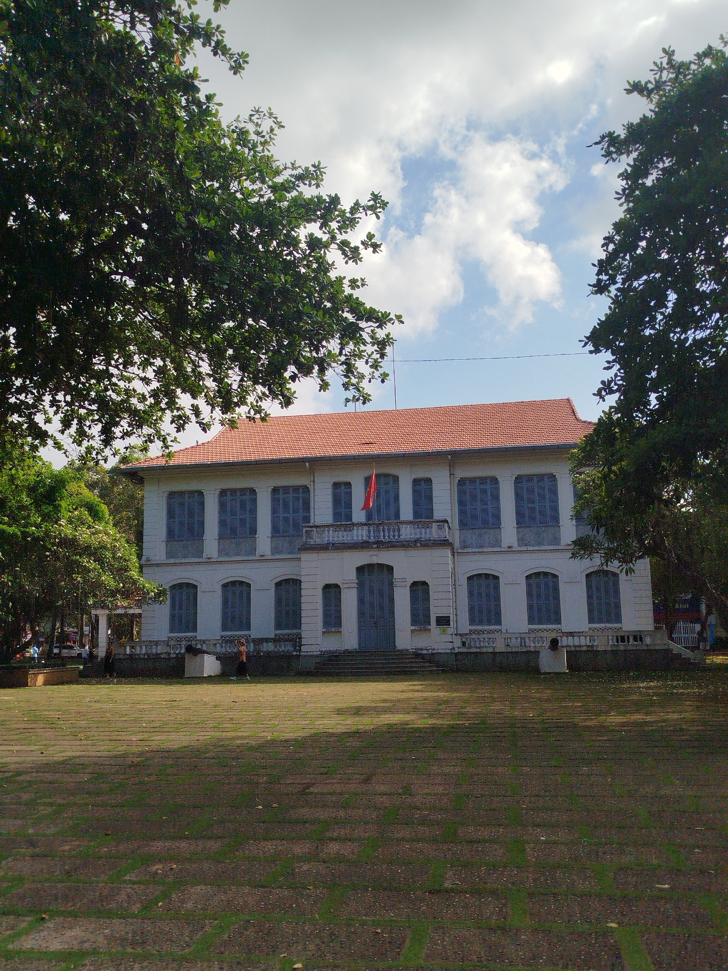 The image shows a two-story white building with a red tile roof in Ho Chi Minh City, Vietnam. Blue shutters cover the windows. A Vietnamese flag hangs from the second story balcony. Trees partially frame the building. The foreground consists of a brick and grass lawn. Several people are visible on the lawn. The sky is cloudy.