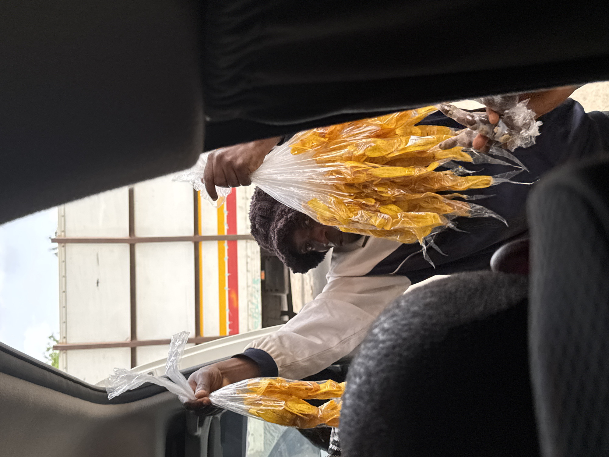 An individual is observed reaching into the open window of a vehicle, offering bags of yellow, chip-like food items wrapped in clear plastic. The person offering the items wears a dark cap, and a light-colored jacket with dark sleeves. The food items appear to be the primary focus of the scene. The image appears to have been taken from inside the vehicle, and the vehicle is parked next to a trailer. The location is the city of Gaga, Nigeria.