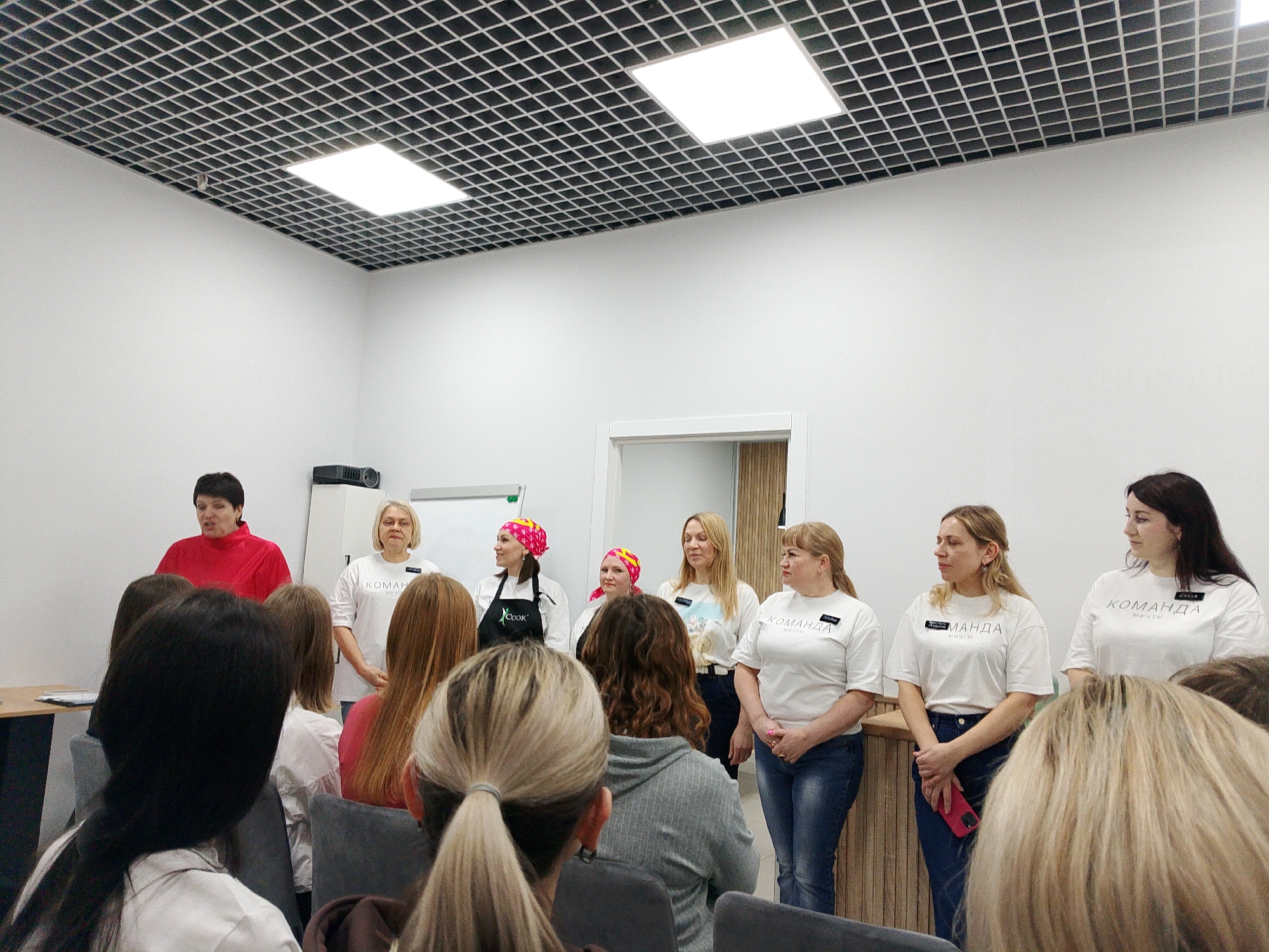 A well-lit indoor scene shows a group of women standing at the front of a room, facing an audience. The setting appears to be in Izhevsk, Russia, although the exact location within the city cannot be determined from the image alone.
The women at the front are dressed in a variety of outfits. One woman is wearing a red top. Several women are wearing white t-shirts with the word "КОМАНДА" ("Team") printed on them, along with a name tag. One woman is wearing a pink headscarf and an apron with the word "COOK" written on it. Most of them are wearing jeans.
The women stand in front of a white wall, with a door visible in the center. A projector is visible on a desk in the corner of the room. The ceiling has a square grid pattern with built-in lighting.
The audience is seated in chairs, facing the group. The backs of their heads and shoulders are visible. The focus is on the front group, suggesting a presentation or a similar event.