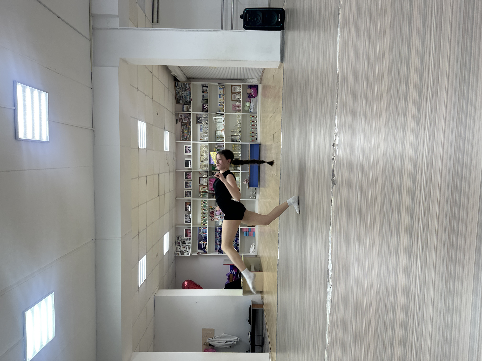 A young woman with a long braid is performing a lunge in a dance studio in Votkinsk, Russia. She is wearing a black leotard and white socks. The studio has a light-colored, striped floor and white walls. A shelving unit filled with various items and trophies is visible in the background. The studio appears to be well-lit with fluorescent lighting.