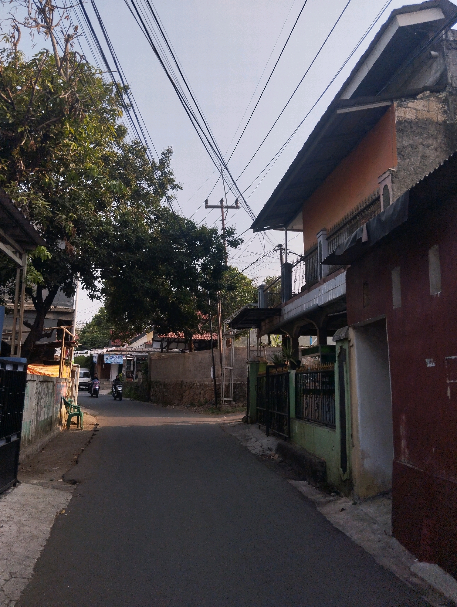 This is a street scene in Bogor, Indonesia. A narrow, paved road curves through a residential area, flanked by houses and trees. Overhead, a dense network of power lines stretches across the sky. On the left, a light-colored wall with graffiti runs alongside the road, with a green plastic chair placed on the sidewalk. A white car is parked further down the road. On the right, a multi-story house with a red lower facade and an orange upper section, featuring balconies and a metal fence, dominates the foreground. Two individuals on motorcycles are visible on the road in the distance, heading towards the viewer. The time appears to be late afternoon or early evening, indicated by the long shadows and the quality of light.