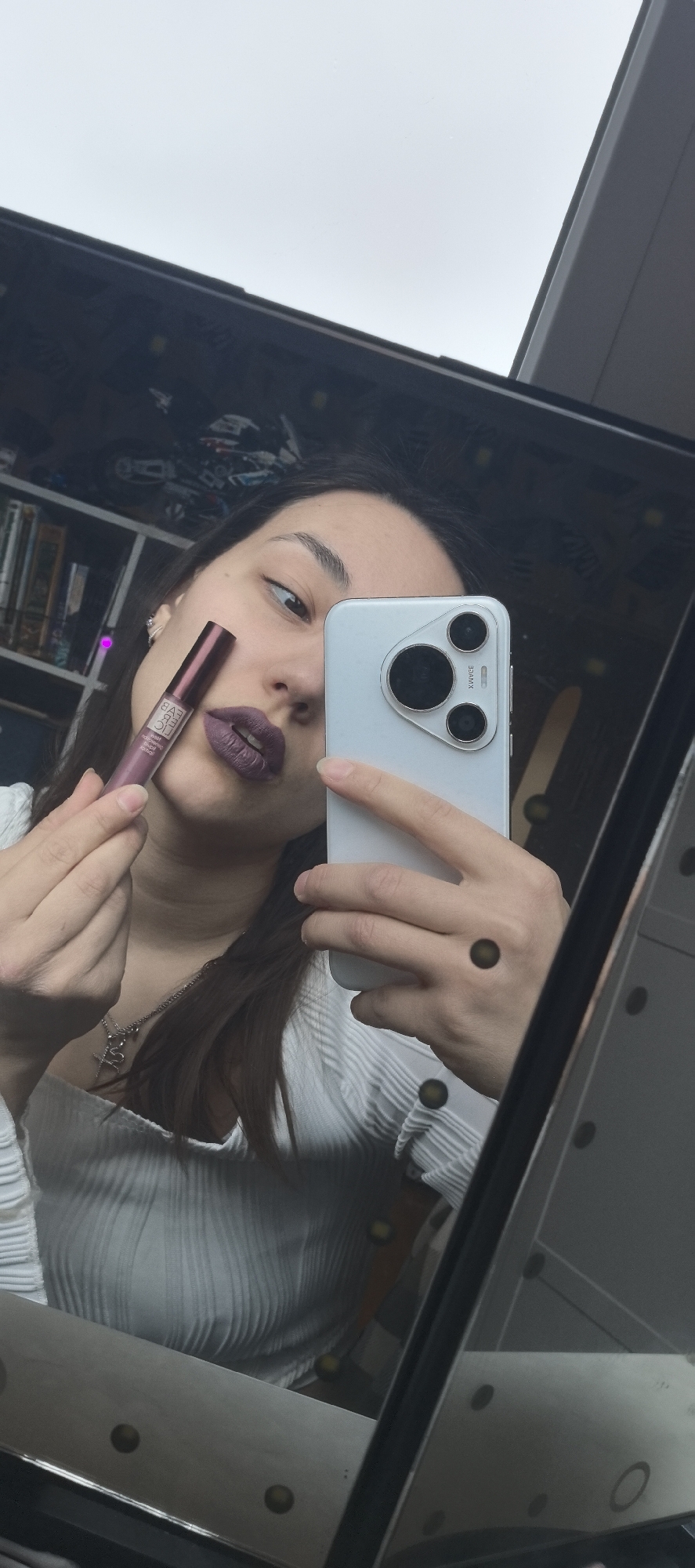 A young woman is captured in a mirror selfie, holding a smartphone in her left hand to take the picture and a tube of liquid lipstick in her right hand. She is looking directly towards the phone's lens with a slightly wide-eyed, playful expression. Her lips are adorned with a dark, matte purple or mauve lipstick, matching the color of the tube she holds.
She has dark hair and is wearing a light-colored, possibly white or off-white, ribbed long-sleeved top. A silver-toned necklace with a pendant, which appears to be a stylized cross or similar symbol, is visible around her neck. Her fingernails are natural and unpainted.
The smartphone in her hand is white or silver, featuring a distinct camera module on the back with three circular lenses and possibly a flash, alongside text that includes "XMWE". The liquid lipstick tube is a similar mauvy-purple shade with a darker cap, and the brand name "SARA" is clearly visible in white capital letters on its side.
The reflection in the mirror reveals elements of an indoor setting behind the woman. To her left, part of a dark bookshelf filled with books and various items is visible. The upper portion of the mirror reflects a bright, possibly overcast sky or a light-colored ceiling. The mirror frame itself has small, dark circular decorative elements along its edges.