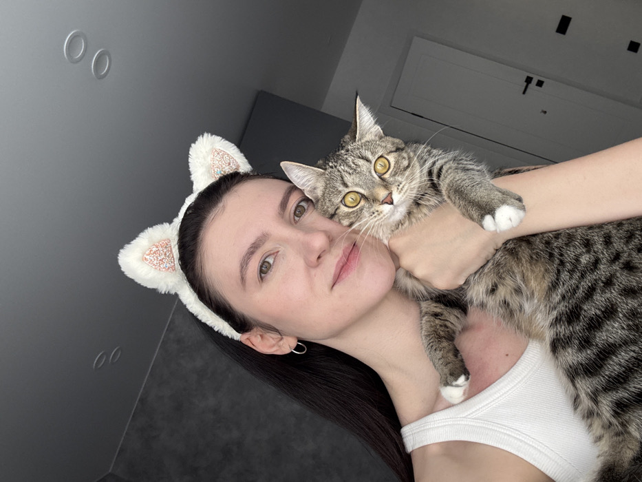 A woman poses for a selfie while holding a tabby cat. She wears a white tank top and a white headband with furry cat ears. The cat has yellow eyes. The background includes a gray wall, two circular indentations on the wall, and an air conditioning unit. The photo is slightly tilted.