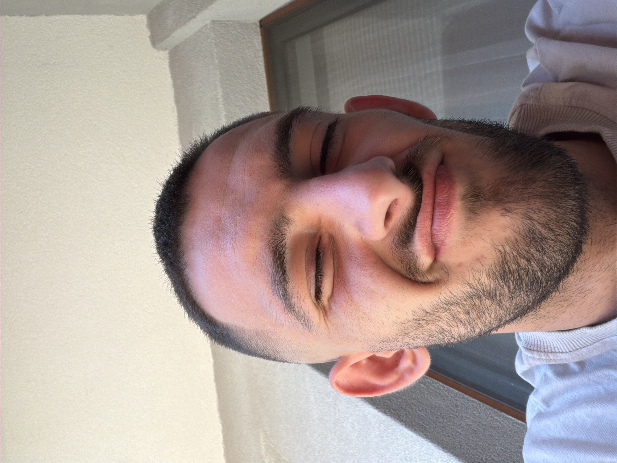 A close-up, slightly angled portrait of a man with a trimmed, dark beard and short, dark hair. His eyes are closed and he has a slight smile. He is wearing a white or light-colored shirt. The background includes a white wall and a window frame with a partially visible window. It is not possible to tell if the image was taken in Plovdiv, Bulgaria, without more context.