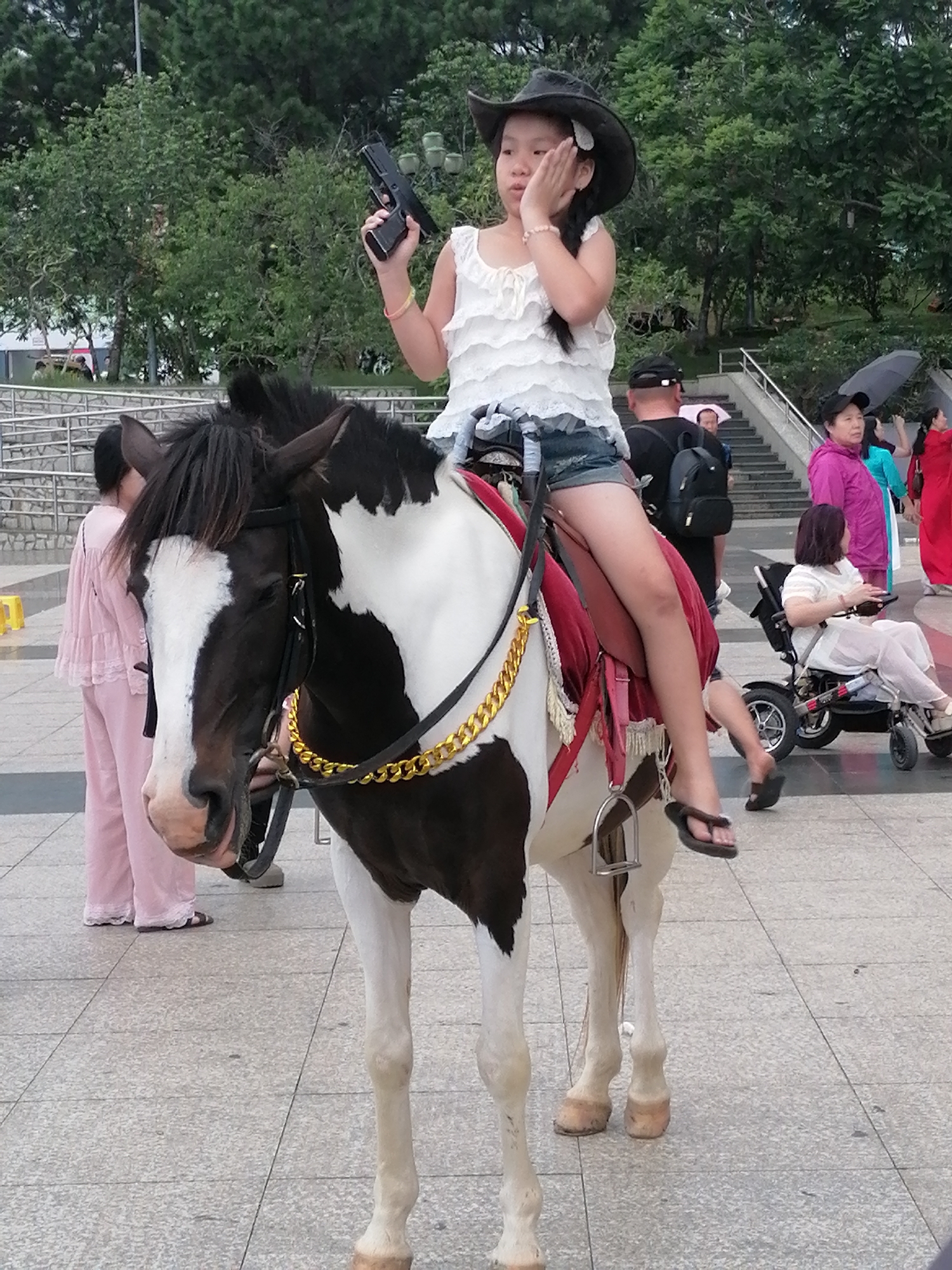 A young girl in a cowboy hat and white ruffled top sits on a black and white horse. She holds a black handgun in her right hand and rests her left hand on her cheek. The horse is adorned with a red saddle blanket and a thick gold chain. In the background, several people are visible, including a woman in a wheelchair, a woman in a pink outfit, and others walking up stairs. The scene takes place on a paved walkway, with trees and foliage in the distance. The overall setting appears to be an outdoor public space, possibly a tourist attraction in Da Lat, Vietnam.