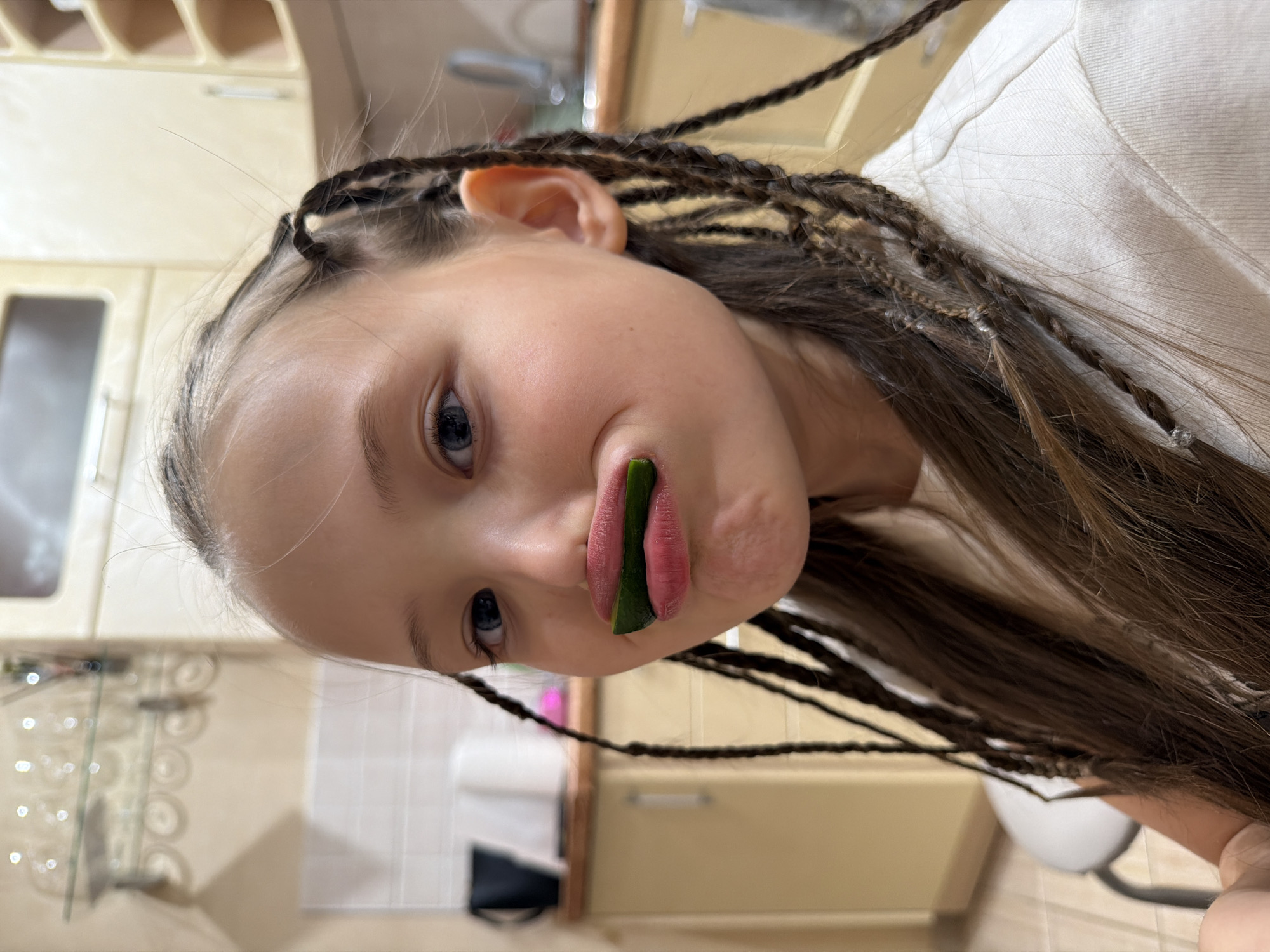 A young girl with brown hair styled in thin braids poses indoors, holding a slice of green vegetable between her upper lip and nose. She has fair skin, light blue eyes, and is wearing a light-colored shirt. The background shows a kitchen with light-colored cabinets and a stainless steel sink. The photograph was taken in a location resembling an interior setting within the city of Chaikovsky, Russia.