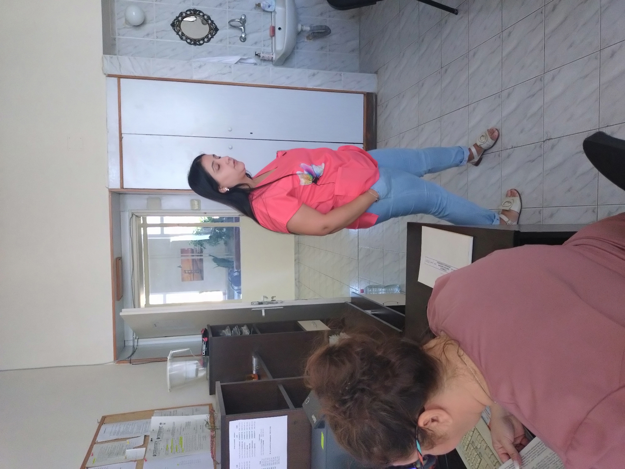 A woman in a coral top and blue jeans lies on a tiled floor in an indoor setting, possibly an office or waiting room in Stara Zagora, Bulgaria. She is adjacent to a cabinet, and her feet are clad in white sandals. A second woman, wearing glasses and a pink shirt, is bent over a desk, seemingly reading or working on paperwork. The desk holds documents and office supplies, and a bulletin board is visible in the background. A bathroom area is also in view with a sink and mirror.