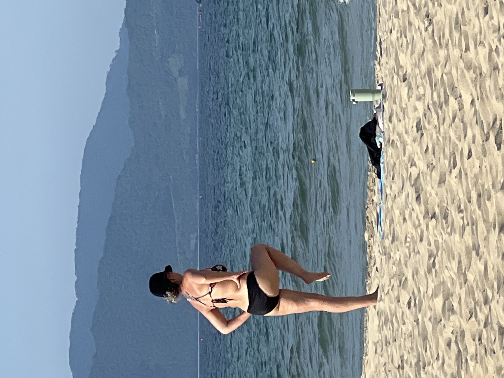 A woman wearing a black baseball cap and a bikini stands on a sandy beach, facing away from the camera. She is in the process of lifting her right leg, bent at the knee, while her left leg remains on the ground. Her right hand is placed on her hip. Behind her, the ocean stretches to the horizon, with distant blue mountains forming a backdrop. On the sand near the water's edge, a black garment, a blue towel, and a green water bottle are visible. The scene appears to be a sunny day. The location is not identifiable as Đà Nẵng, Vietnam, based on visual information alone.