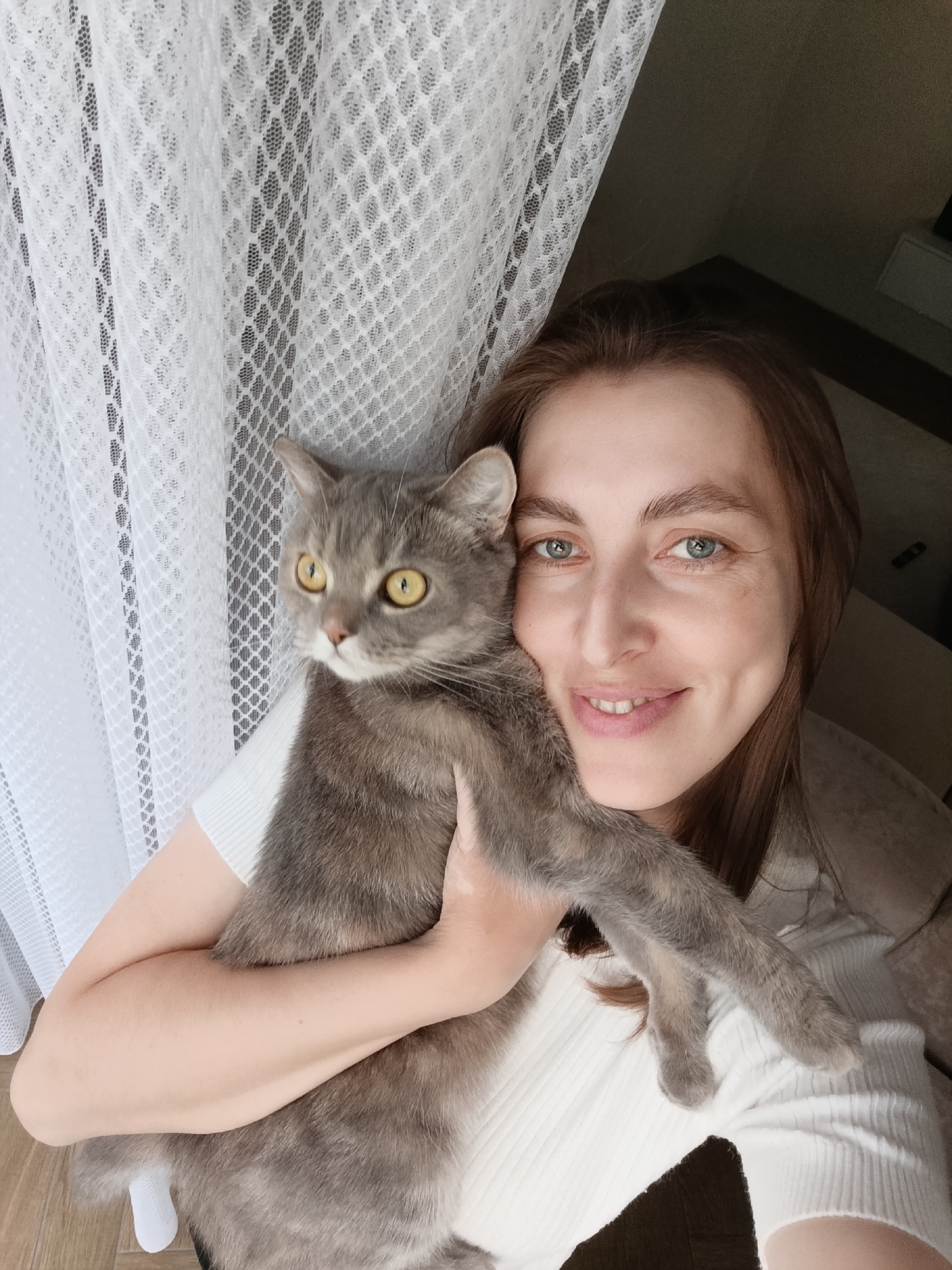 A woman is holding a grey cat in front of a white lace curtain and a dark wall. The woman is looking at the camera and smiling. The cat is looking to the left. The scene appears to be indoors, possibly in a residential setting. There are no identifiable landmarks or clues to pinpoint the location in Chaikovsky, Russia.