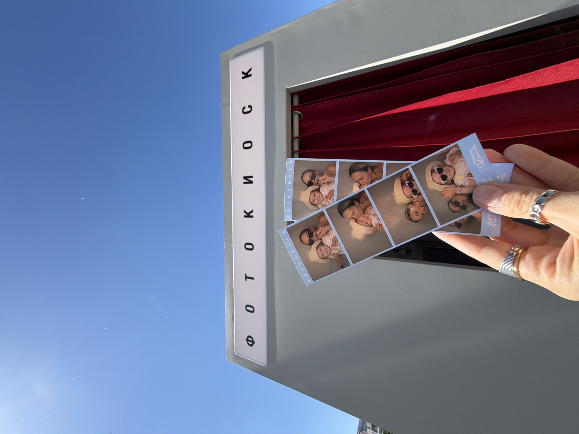 A right hand, adorned with a two-tone ring on the ring finger and a silver multi-circle ring on the index finger, holds two vertical photo booth strips. Each strip contains multiple square photographs depicting an adult woman with sunglasses and a young child wearing a hat and heart-shaped sunglasses, posing together with expressions of joy and affection. The strips include the text "ПАРКИ МОСКВЫ" (Parks of Moscow) and "ФОТОКИОСК" (Photo Kiosk). In the midground, a grey, angular photo booth structure is visible, featuring a white sign above its entrance with the black Cyrillic text "ФОТОКИОСК." A rich, dark red pleated curtain covers the booth's entrance, slightly open on the right. The background consists of a clear, bright blue sky devoid of clouds. The scene is located in Khimki, Russia.