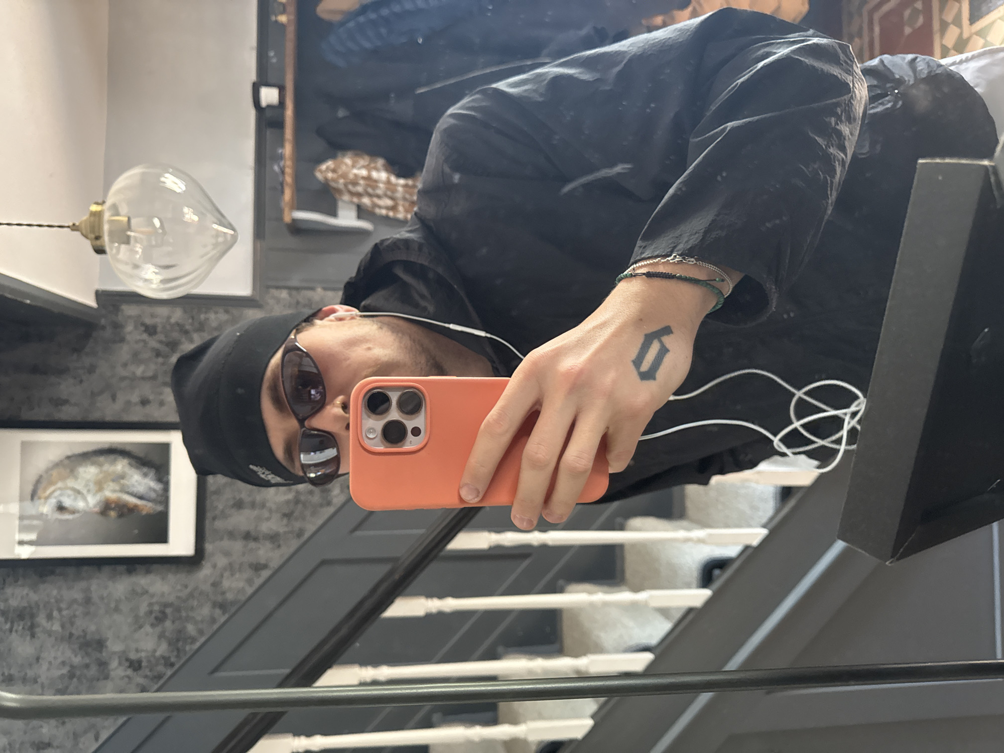 A reflection captured in an indoor setting, likely in Stotfold, United Kingdom, shows a person taking a selfie. The person, appearing to be male, is seen from the chest up, reflected as if lying on their side due to the camera's orientation. They are wearing a black beanie, dark sunglasses, and a dark-colored jacket or shirt, possibly made of a textured, lightweight fabric. White wired earbuds are visible, with one extending into the person's ear.
The person is holding an orange iPhone, likely a Pro model, with its triple camera setup visible on the back. On the back of their left hand, a black tattoo in a Gothic-style font is present, resembling the letter 'D'. Several bracelets adorn the wrist, including a black beaded one, a silver chain, and a green beaded one.
The background, also reflected, reveals an interior space with a distinct aesthetic. To the left, a unique light fixture hangs, featuring a clear, teardrop-shaped glass bulb with internal elements visible. Below it, a framed picture hangs on a textured gray wall, depicting what appears to be an abstract or animal form, possibly a marine creature like a seal or walrus. A staircase with dark gray risers and white balusters is prominent in the lower half of the reflection, with a dark handrail running along it. A partially obscured doorway or cabinet is also visible in the upper right. The overall activity is a self-portrait captured via a reflective surface.