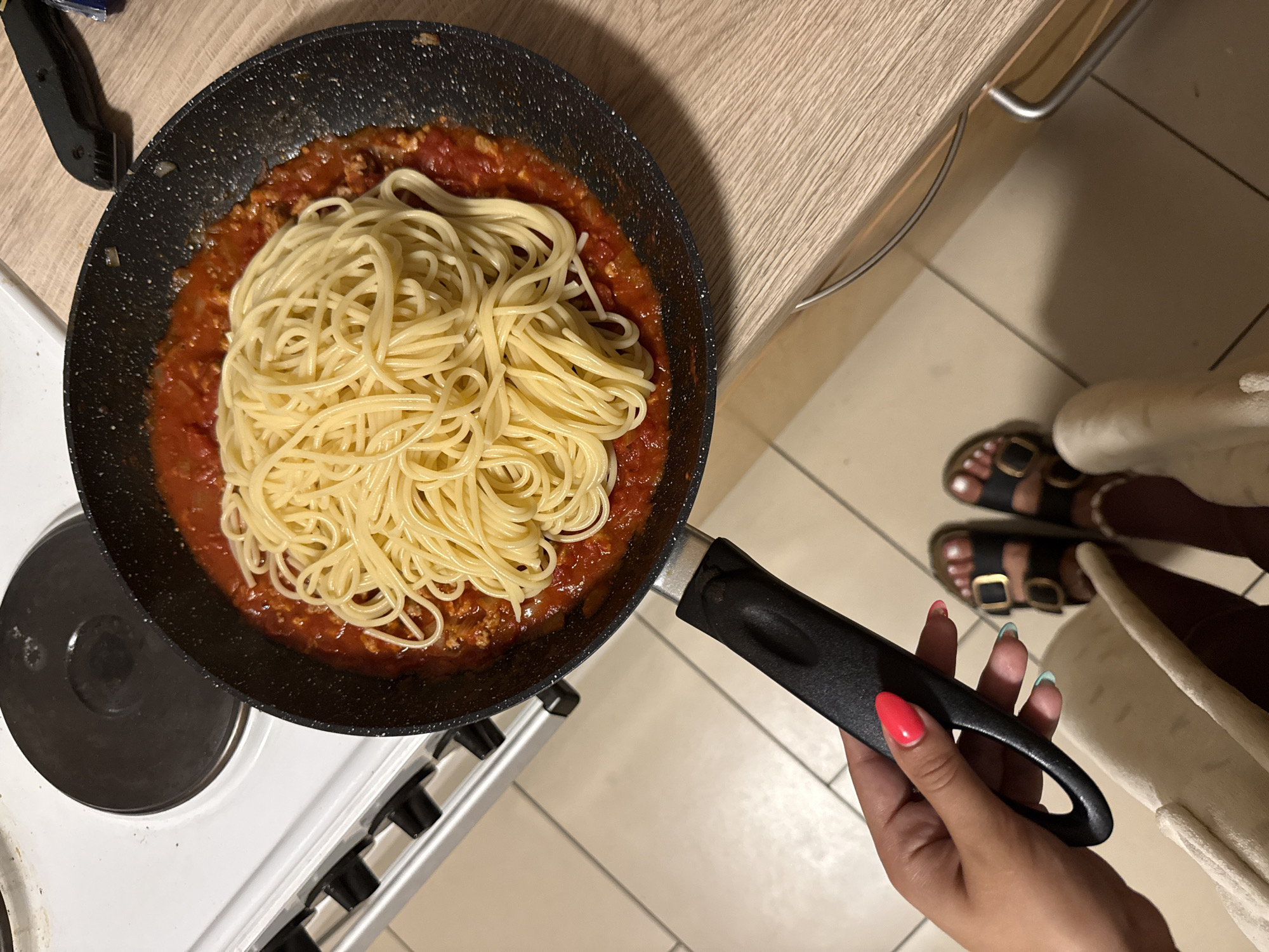 A person's hand with bright pink fingernails is holding the handle of a black frying pan filled with spaghetti and tomato sauce. The pan is on a stovetop next to a kitchen counter. The person is wearing black sandals. The scene appears to be a kitchen in a home.