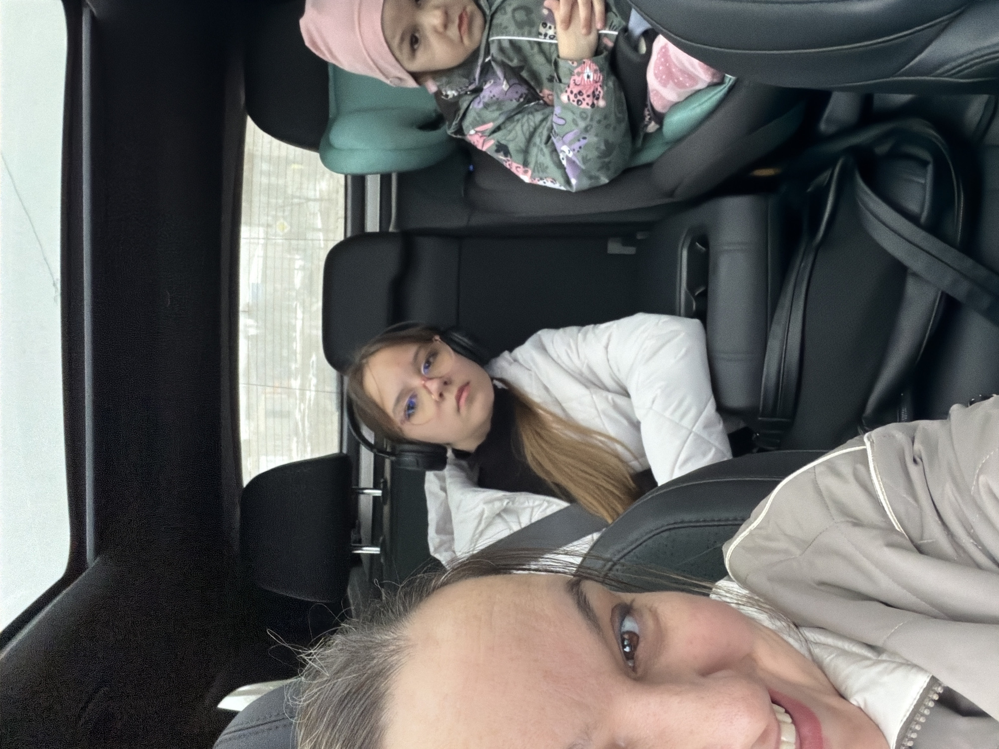The image shows three people in a car. In the front seat is a woman with short brown hair and a beige jacket taking a selfie. In the back seat is a young woman with glasses and headphones. Behind the young woman is a toddler in a car seat wearing a pink hat and a printed jacket. The car appears to be stopped at a traffic light or in slow traffic, and the setting suggests it may be in Марковский, Russia, due to the user’s prompt.