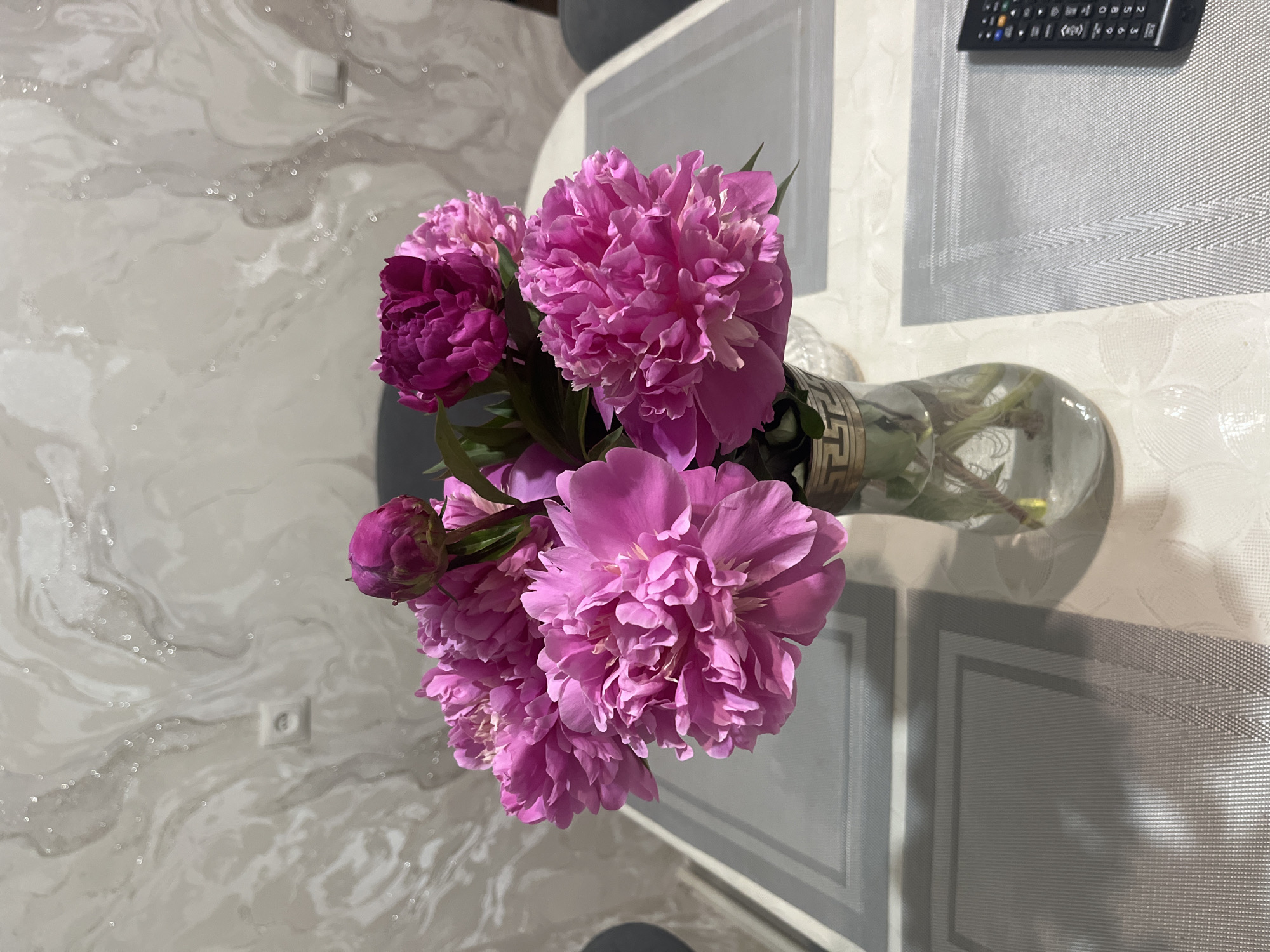 A high-angle shot displays a bouquet of pink peonies in a glass vase. The flowers, varying in shades of pink and in stages of bloom, dominate the central part of the frame. The vase, adorned with a Greek key design at the top, appears to contain water, with some stems visible.
The bouquet rests on a surface with a light-colored, textured tablecloth. A remote control is partially visible in the upper right corner. The background includes a wall with a marbled pattern and an electrical outlet. The scene is likely indoors. There's no information to verify that it is located in Kaspiysk, Russia.