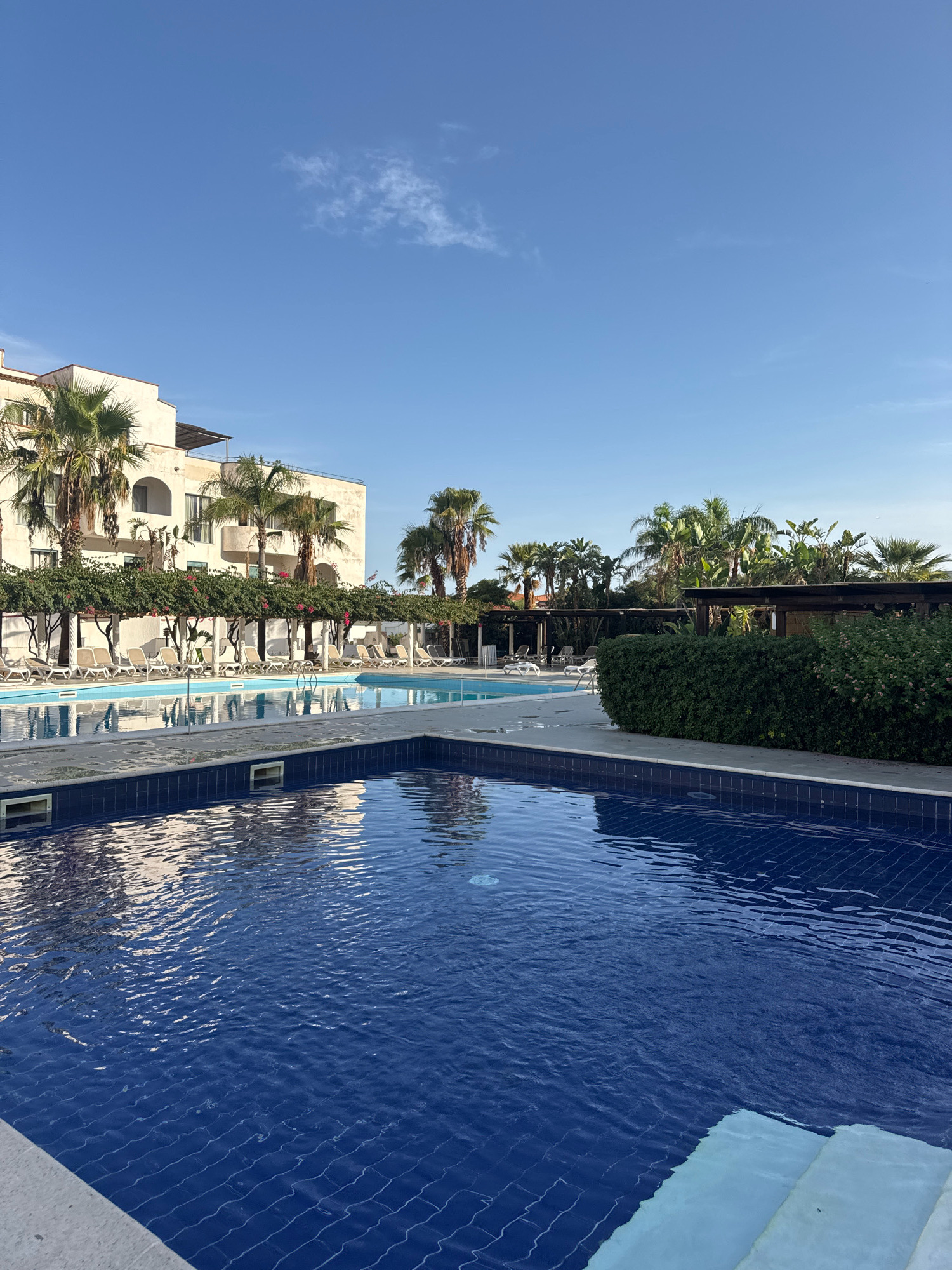 A bright, sunny day illuminates a resort scene in Isola delle Femmine, Italy. In the foreground, a pristine, blue swimming pool with tiled edges reflects the surrounding environment. The water is calm with subtle ripples. Steps are visible at the bottom right corner of the pool.
Beyond the pool, a paved area is lined with rows of empty lounge chairs under a covered walkway supported by white columns and adorned with green foliage and pink flowers. Behind the lounge chairs, a multi-story building with a light-colored facade and arched windows stands prominently, interspersed with tall palm trees.
Further back, another section of the swimming pool is visible, alongside lush green bushes and trees, including more palm trees and tropical foliage. Wooden pergolas are scattered throughout the area, some covered with greenery, suggesting shaded seating or relaxation spots. The sky above is a clear, bright blue with a few wispy white clouds. The overall impression is one of a tranquil and well-maintained resort, perfect for a vacation.