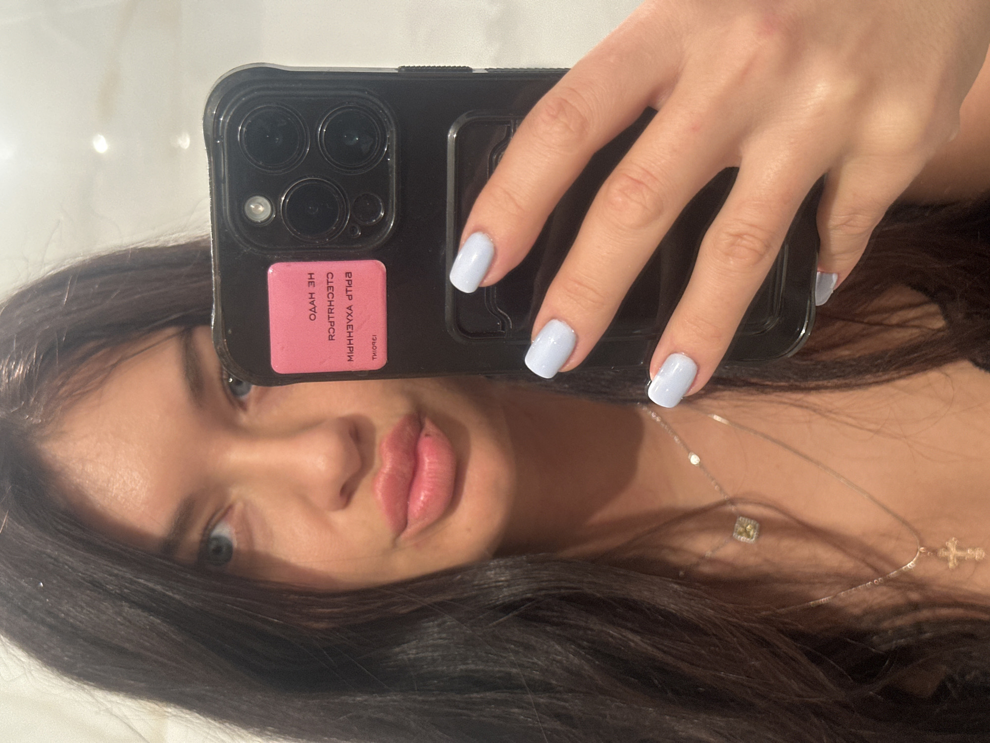 A mirror selfie captures a young woman, partially visible, holding a smartphone. The image is taken indoors, likely in a well-lit room or bathroom, against a light, plain background. The woman, located in Moscow, Russia, has dark, flowing hair and bright blue eyes, which are prominently featured. Her lips are slightly parted in a soft pout. She has a light skin tone.
Her right hand, adorned with light blue or periwinkle nail polish, holds a black, possibly rugged, smartphone case. The phone appears to be an iPhone, evident from its distinct triple-camera array and flash. On the back of the phone case, there is a rectangular pink sticker. Due to the mirror reflection, the text on this sticker is reversed. Partially decipherable text includes the Russian phrase "НЕ НАДО" (NE NADO), which translates to "don't need" or "don't have to." Other characters on the sticker are stylized and difficult to fully interpret when reversed.
The woman wears two layered necklaces. The upper necklace features a small, subtle square-shaped pendant, while the lower necklace has a small cross pendant. The overall mood is casual and personal, characteristic of a self-portrait shared on social media.