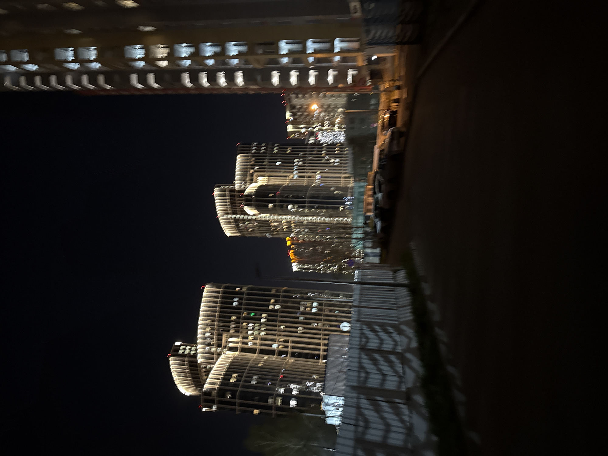 The image shows a night view of buildings in Khimki, Russia, likely taken while in motion, resulting in motion blur. Three high-rise buildings are visible, illuminated with warm lights emanating from numerous windows, suggesting residential occupancy. The buildings feature a modern architectural style, with a tiered design on some. The foreground is dark, with what appears to be a road or pathway along the right side. Faint vegetation is also visible in the lower right. Another building with similar lighting is at the top left of the image. The overall scene appears to be a nighttime urban setting with a focus on the illuminated buildings.