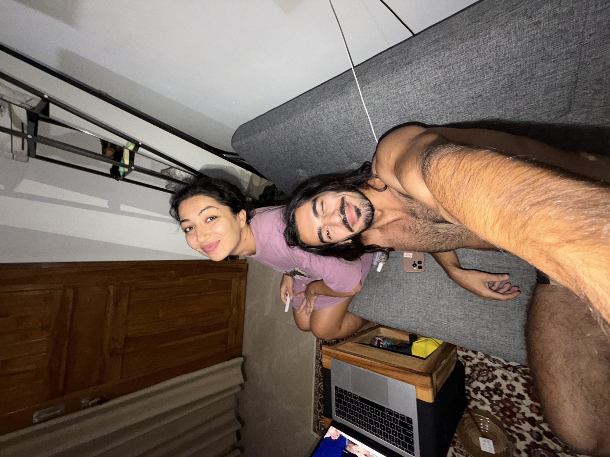 An overhead selfie captures a man and a woman relaxing indoors, likely in North Kuta, Indonesia. The man, shirtless with long dark hair and a beard, is in the foreground, smiling broadly at the camera, seemingly taking the photo. Behind him, a woman with dark hair, wearing a light purple top, also smiles at the camera, holding a white stick-like object.
They appear to be reclining on or beside a grey upholstered sofa or bed. To their left, a wooden bed frame or headboard is visible, and above it, a black metal shelving unit is mounted on the wall. A white cord hangs from the ceiling near the sofa. Adjacent to the man, a wooden side table holds a silver laptop and a small yellow container. A light-colored smartphone with two cameras rests on the grey fabric surface near the man's hand, alongside a clear bottle. A patterned rug covers the floor beneath the table. Another screen, possibly a tablet or second laptop, displaying images, is partially visible in the bottom left corner. The indoor lighting suggests it is evening or night, and both individuals convey a relaxed and happy mood.