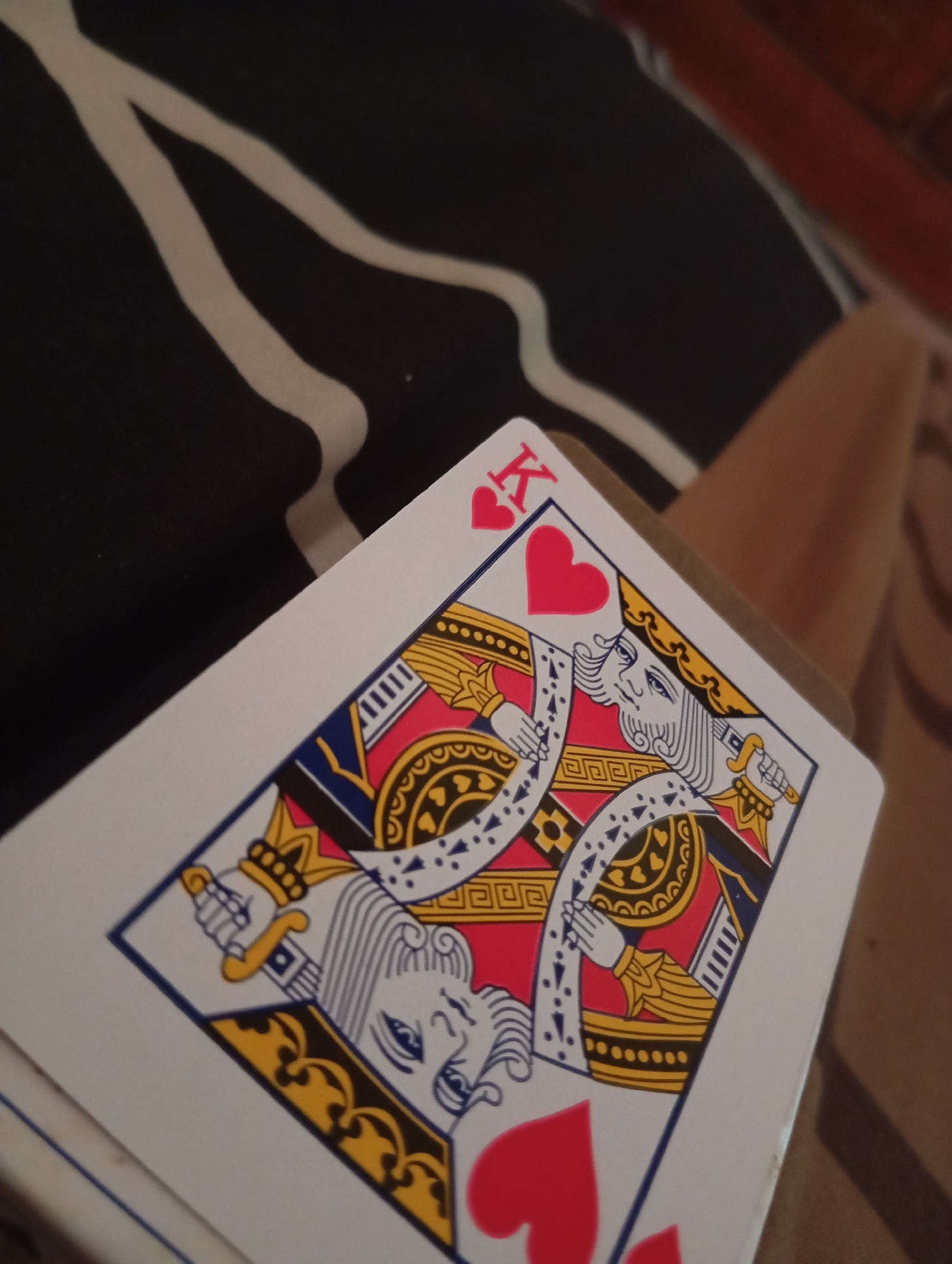 A close-up, angled shot features the face of a King of Hearts playing card. The card is partially obscured by other cards beneath it and is angled to the right. The "K" and a heart symbol are visible in the top left corner. The central design of the card depicts a king with a crown and a scepter in his hand, rendered in red, blue, and yellow. The background consists of a dark surface with white wavy stripes and a blurred reddish-brown object. No city or location information is discernible from the image itself.