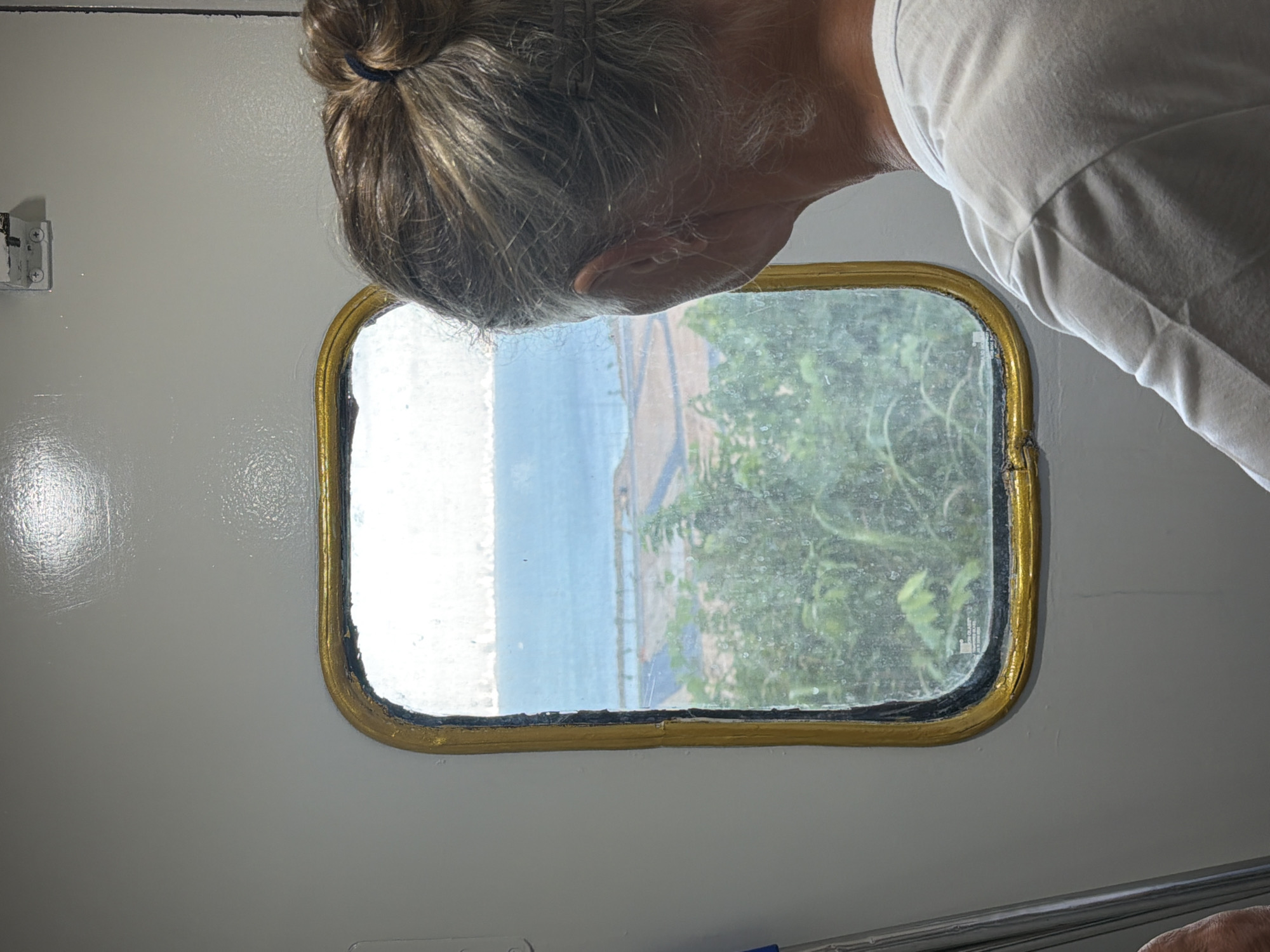A person with graying hair styled in a bun, wearing a white t-shirt, is looking out of a small, rectangular window with rounded corners and a gold frame. The window offers a view of what appears to be the coast, with a light blue body of water, a strip of land, and green vegetation. The window is set into a light-colored, possibly white, surface. The image is taken indoors, possibly inside a vehicle or structure near the coast. Based on the hints, the city of Đà Nẵng, Vietnam, is relevant to the location.