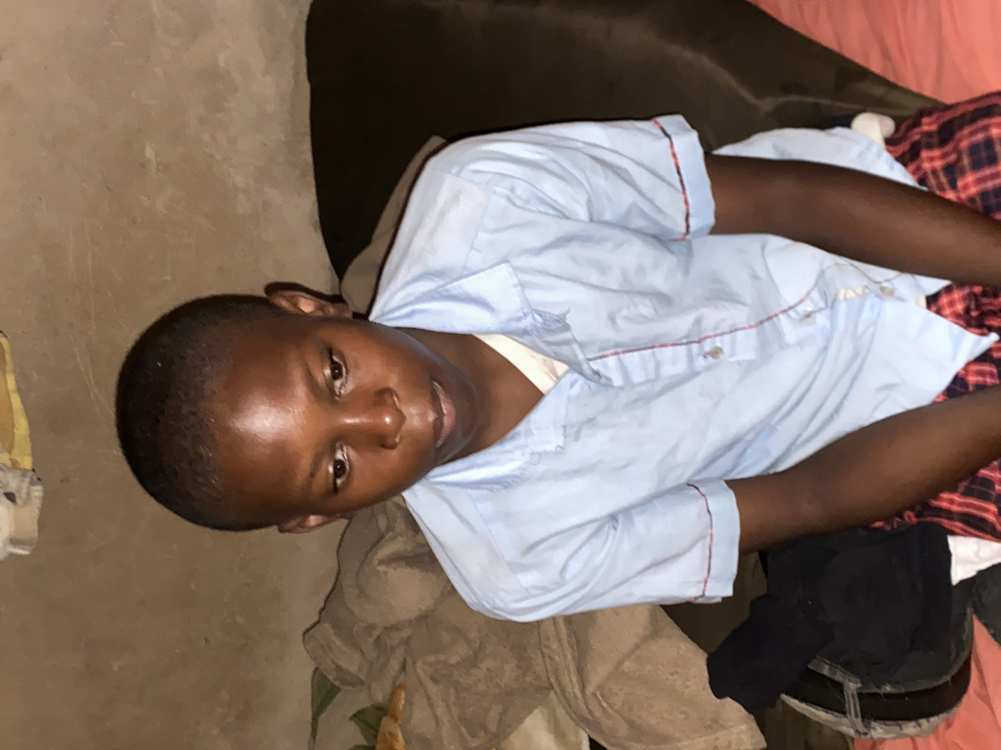 A young African male is lying on a bed. He has short, dark hair and is wearing a light blue shirt with red trim. He's resting on a brown sofa, and a red plaid blanket is next to him. The background includes a cream-colored wall and some discarded clothes with a visible belt. The location appears to be indoors, though specific information about Bukuru, Nigeria is unavailable from the visual content alone. The scene portrays a casual, relaxed moment.