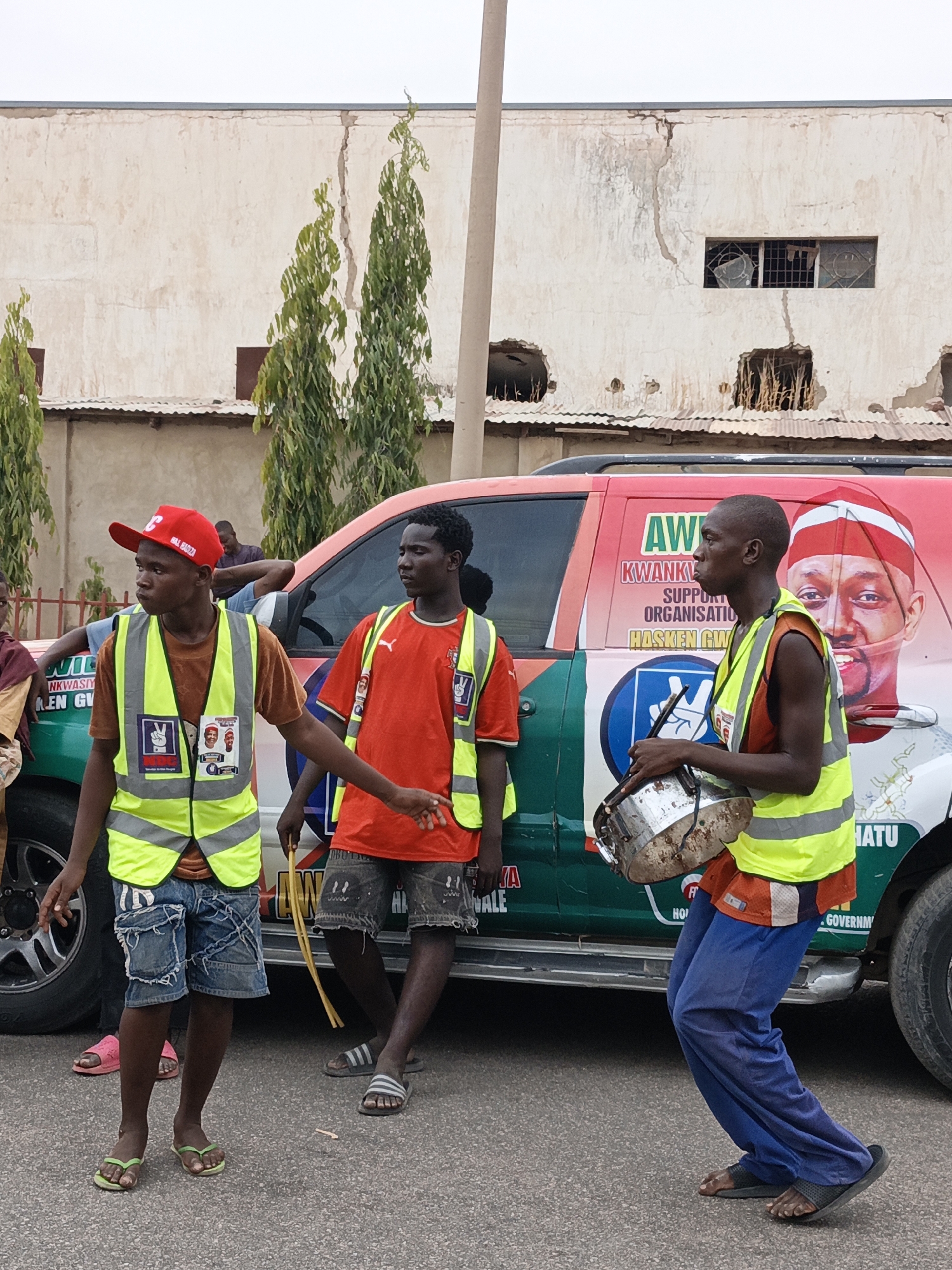 In a city street scene, likely Tudun Wada, Nigeria, three young men stand next to a vehicle plastered with political endorsements. One man, wearing a red cap and yellow reflective vest over a brown shirt, has denim shorts and green sandals. Another, in an orange Puma shirt and reflective vest, wears grey shorts and striped sandals. The third, playing a small, worn drum, wears a reflective vest over an orange shirt and blue pants with black sandals. The vehicle behind them is green, white, and red, with a large portrait of a man, presumably a politician. Text on the vehicle includes "AW", "KWANK", "SUPPORT ORGANIZATION", "HASKEN GW", and "HATU". In the background is a two-story building with a crumbling facade. Three tall, thin trees line the street, and a utility pole is visible. The road is paved.