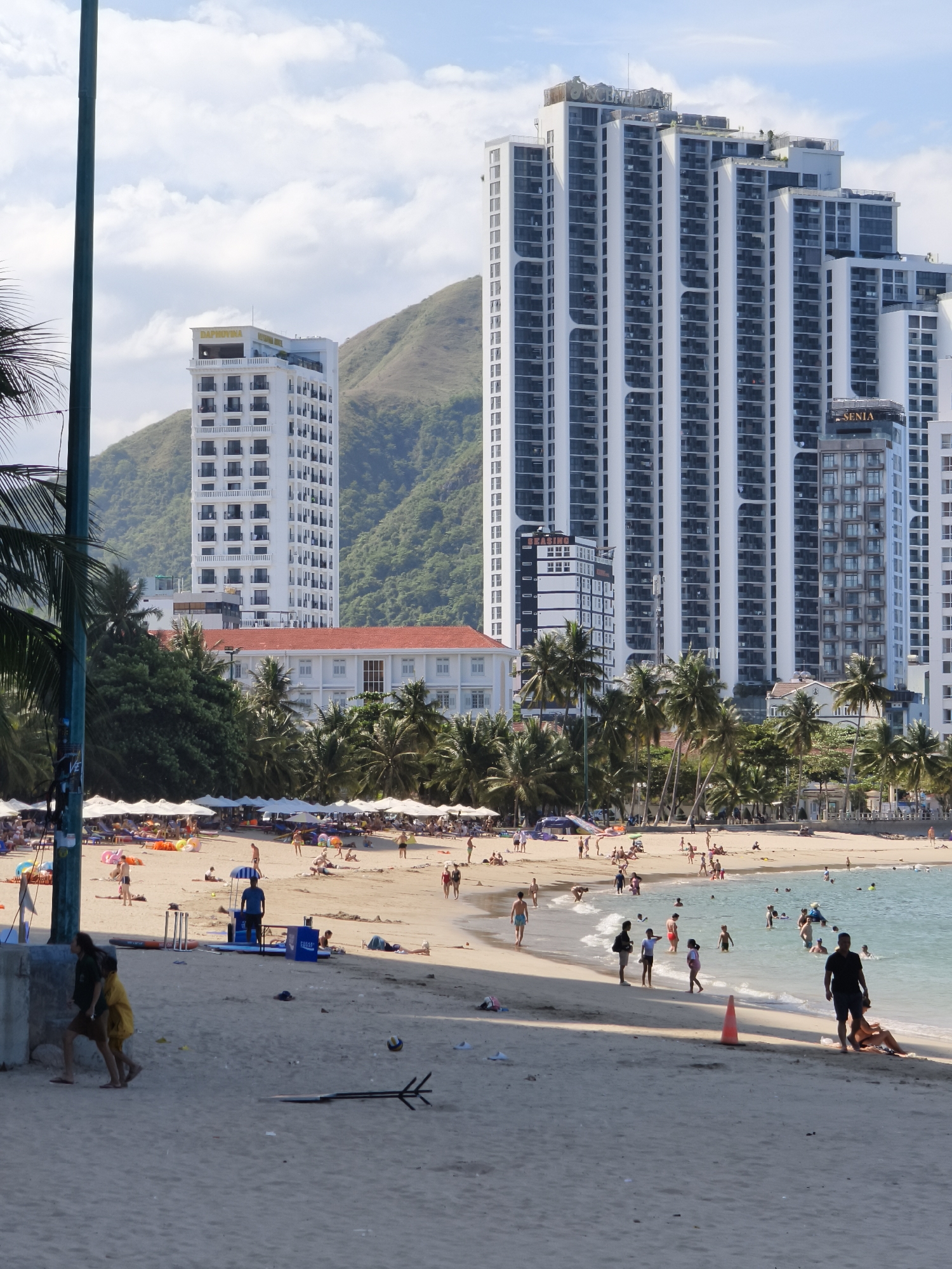 A bustling beach scene unfolds in Nha Trang, Vietnam, under a partly cloudy sky. In the foreground, two children walk across the sand, one wearing a yellow dress and the other a green top. A volleyball lies on the sand, near a black, flat object with prongs. In the middle ground, a lifeguard stands near a blue cooler and surfboards, while beachgoers lounge under white umbrellas. Palm trees line the shore, separating the beach from a row of buildings. Dominating the background are several modern, multi-story buildings, some with distinctive wavy facades. In the distance, a lush green mountain rises, its slopes covered in foliage. The turquoise water of the ocean is dotted with swimmers and a few small boats.