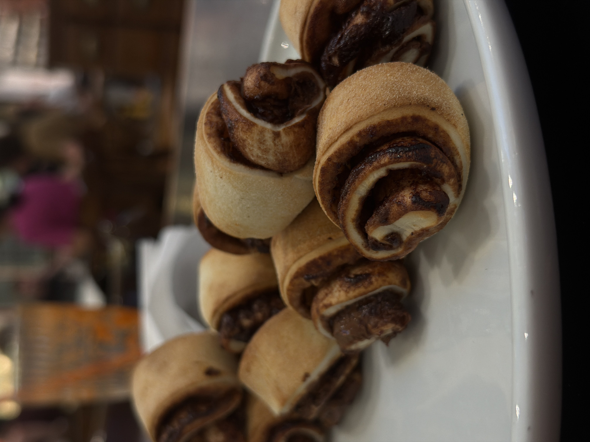 A close-up, eye-level shot focuses on a pile of freshly baked rugelach pastries on a white plate. The pastries are arranged in a rustic, organic cluster, with some overlapping and others slightly separated. Each rugelach features a swirled interior of chocolate filling, visible through the golden-brown, flaky dough. The dough has a slightly porous texture, indicating a soft, yielding crumb. The lighting is soft and natural, casting subtle shadows that highlight the contours of the pastries. The background is blurred and indistinct, showing hints of a restaurant or cafe environment. The overall impression is one of warmth and comfort, with the focus solely on the inviting appearance of the rugelach. It is plausible that this image was taken at a cafe or bakery in Ramat Gan, Israel, given the popularity of rugelach in Israeli cuisine.