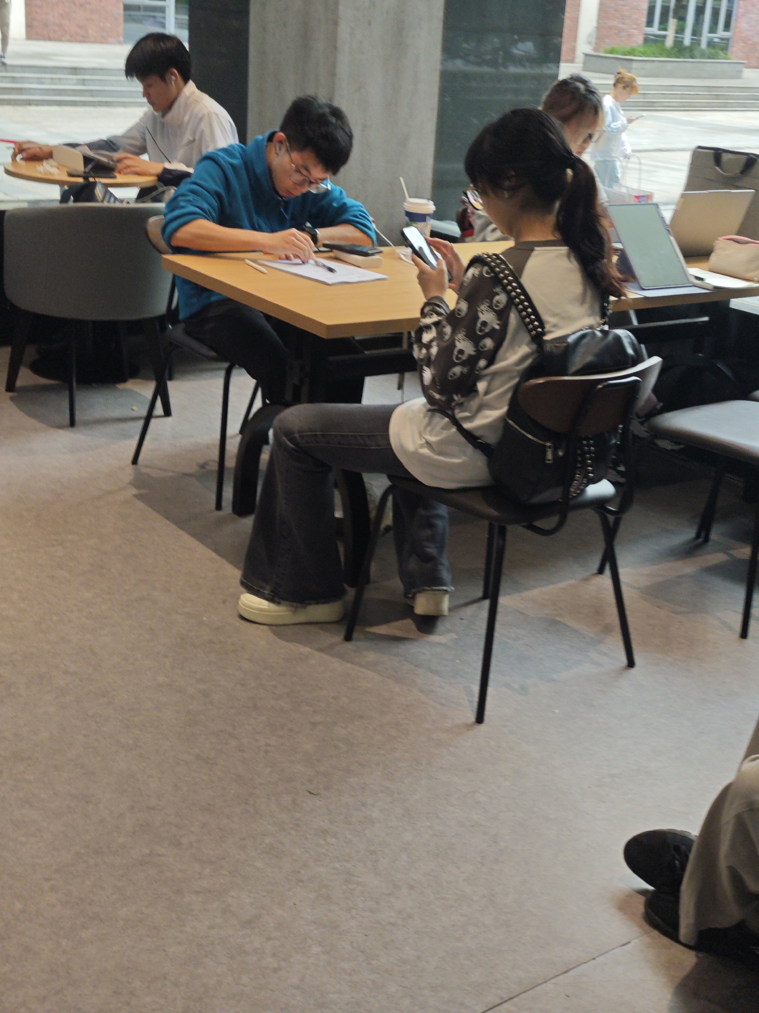 In a brightly lit indoor space, possibly a cafe or study area, a group of young adults are engaged in various activities. In the foreground, a woman with dark hair tied in a ponytail is seated, looking at her phone, which she holds in her right hand. She is wearing a white top with a dark, patterned design and a black backpack. To her left, a man in a blue hoodie and glasses is bent over a table, appearing to write on a piece of paper, with a laptop and other items nearby. Further back, another man in a white shirt is seated at a table with a laptop, and a woman with blonde hair stands in the background near some stairs. The environment features light-colored flooring and wooden tables. The scene appears to be a contemporary public space conducive to study or casual work.