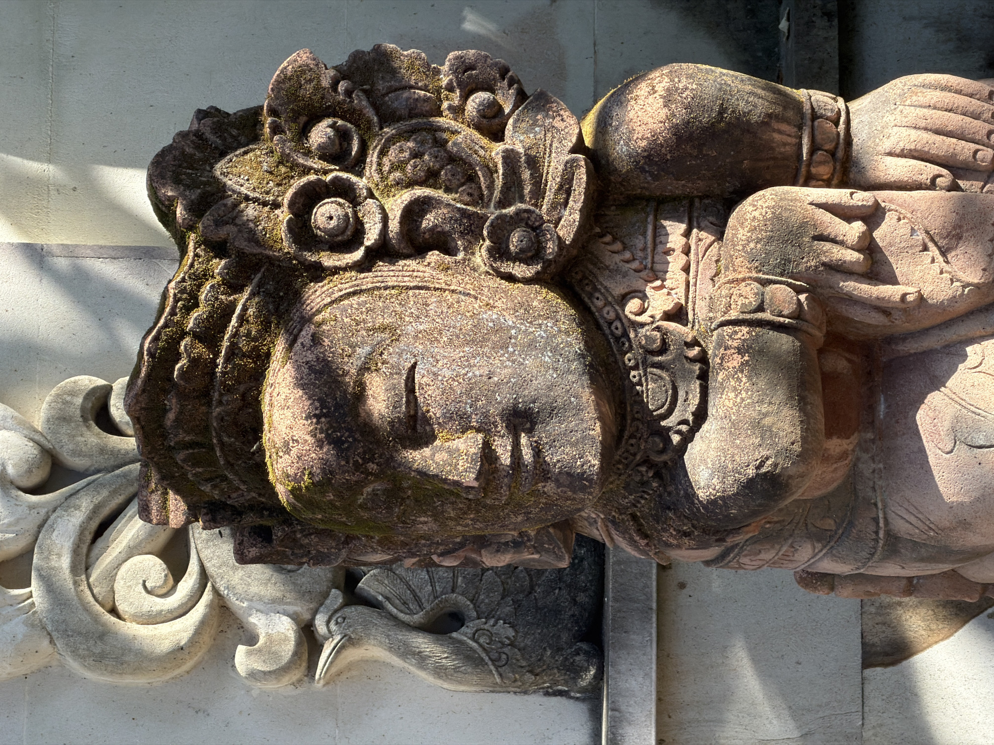The image displays a stone statue of a figure with intricate headdress and jewelry, positioned in what appears to be an outdoor setting. The statue is partially covered in moss, indicating age and exposure to the elements. Behind the statue, a relief carving of a mythical bird and swirling decorative elements are visible on a textured wall. Sunlight casts shadows on the wall and the statue, suggesting it is daytime. The statue is located within the Pura Besakih temple complex in Denpasar, Indonesia.