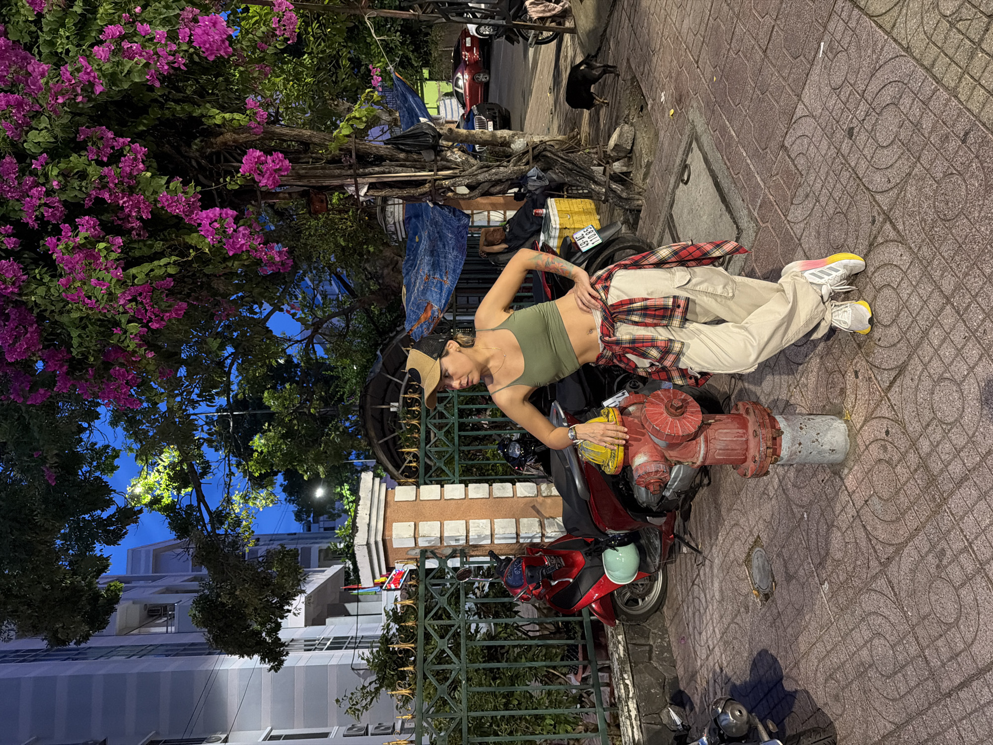 A woman stands on a tiled sidewalk in Nha Trang, Vietnam, leaning against a red fire hydrant. She wears a green tank top, tan cargo pants, a red and black plaid shirt tied around her waist, and white sneakers with yellow and pink accents. A tan baseball cap is on her head. To her left, a red scooter is parked. In the background, a building with balconies and air conditioning units is visible, along with trees bearing bright pink flowers. A blue tarp is stretched over some objects behind a tree. A small black dog stands further down the sidewalk. Several cars are parked on the street in the distance, and a person is seen sleeping on a yellow cooler next to a motorcycle. The sky is dark blue, indicating it is either dusk or nighttime.