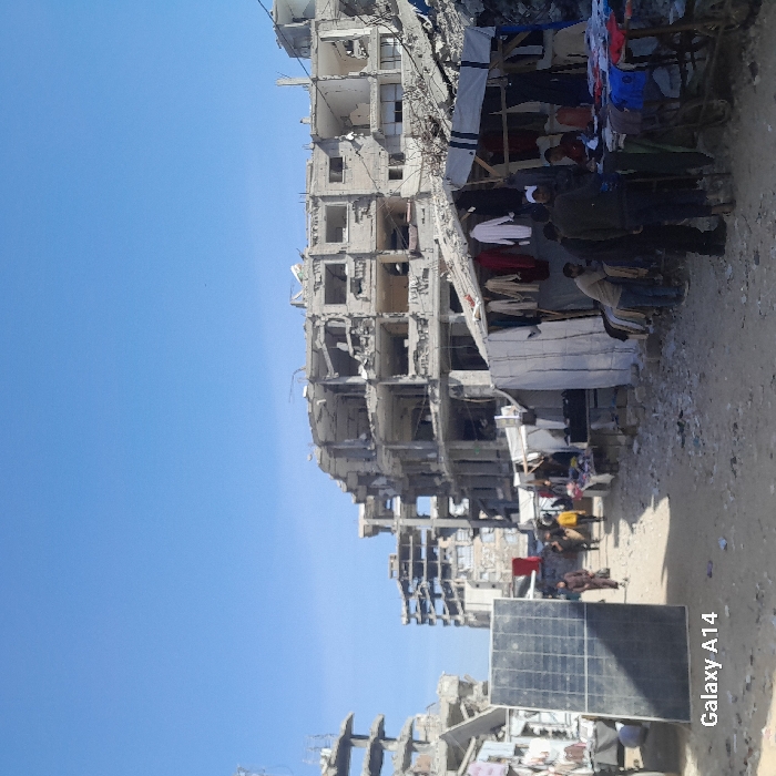 This image depicts a wide, unpaved path in Khan Yunis, Palestinian Territory, flanked by extensively damaged multi-story concrete buildings under a clear blue sky. The structures, primarily in the background, show significant destruction with blown-out facades, exposed rebar, and rubble. In the midground and foreground, a makeshift market operates along the right side of the path. Several stalls are constructed from tarpaulins and sheets, displaying clothes, including jackets, shirts, and traditional garments, hanging for sale. Multiple individuals are present: several men are gathered around the market stalls, with one man in a dark jacket leaning over a cart laden with clothes. Further down the path, a few figures, including women, are seen walking away from the viewer. On the left side of the path, a large, rectangular black solar panel stands upright. The ground is dusty and strewn with debris. Text in the bottom-left corner identifies the camera as "Galaxy A14."
